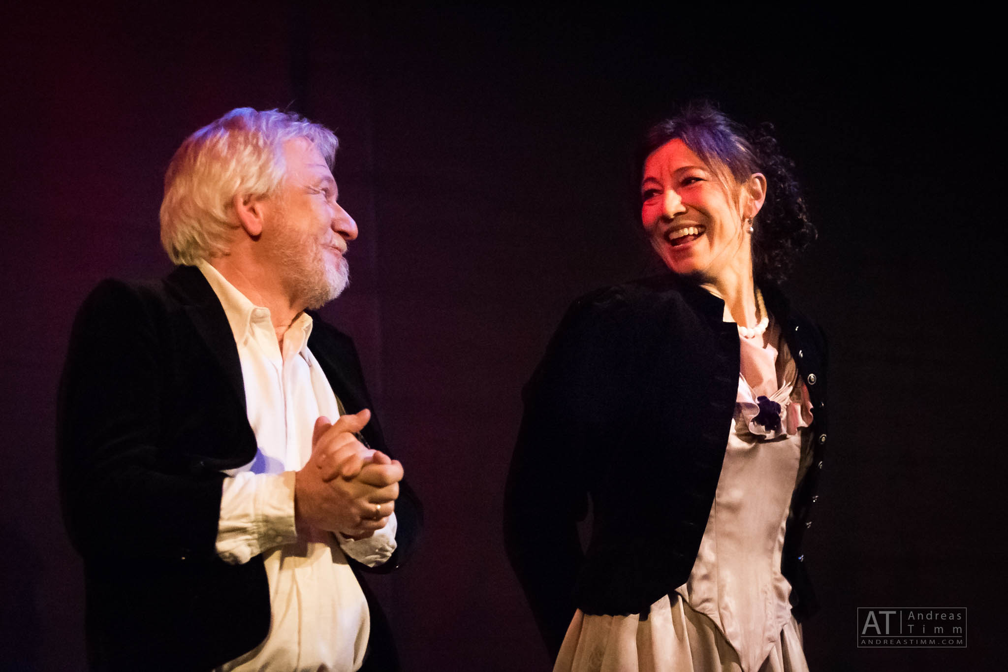 Two actors in period costumes share a joyful moment on stage, warmly lit in a theatrical setting.