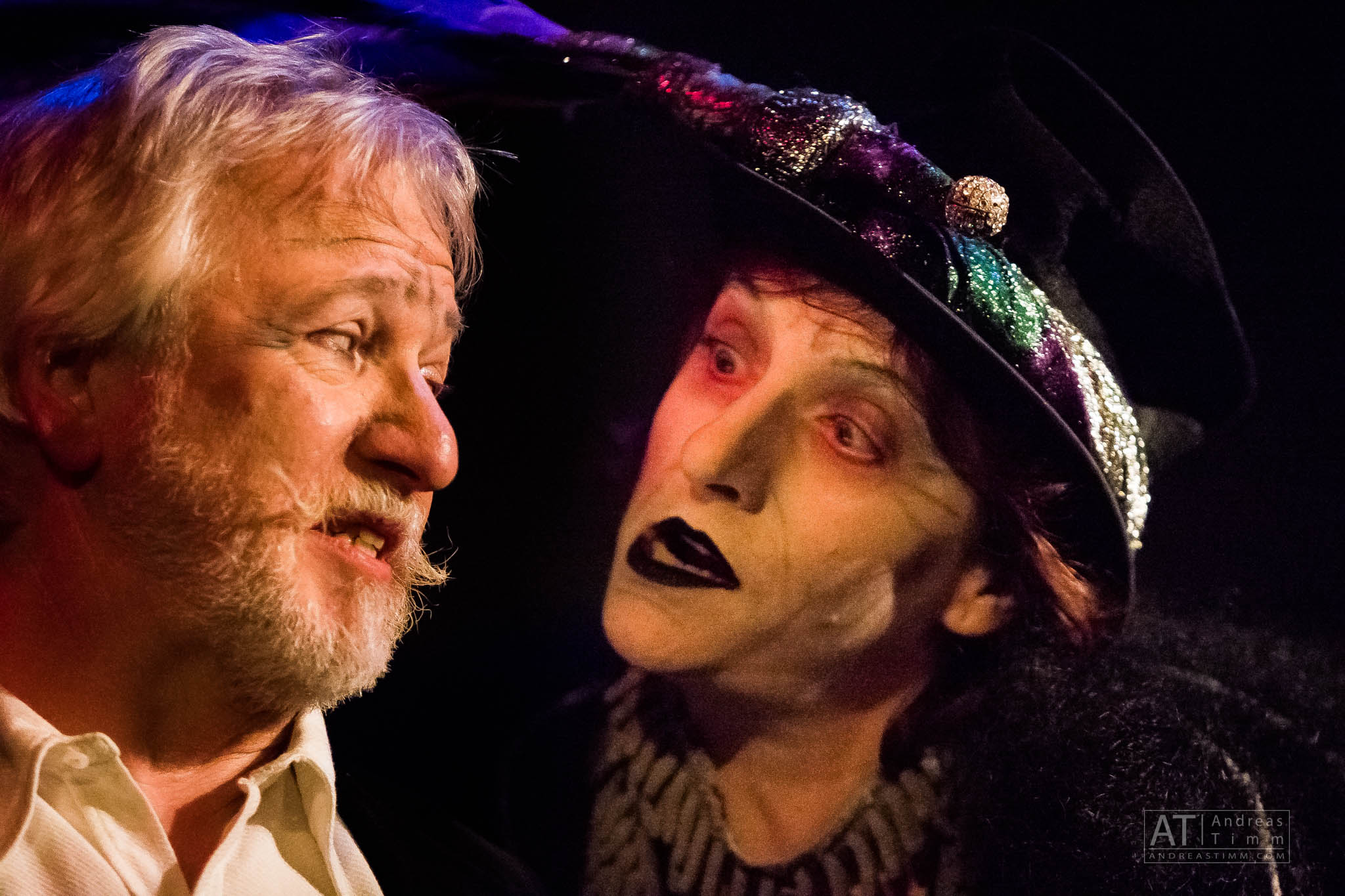 Elderly man speaking to person in colorful witch-like costume and makeup, dramatic theater performance scene.