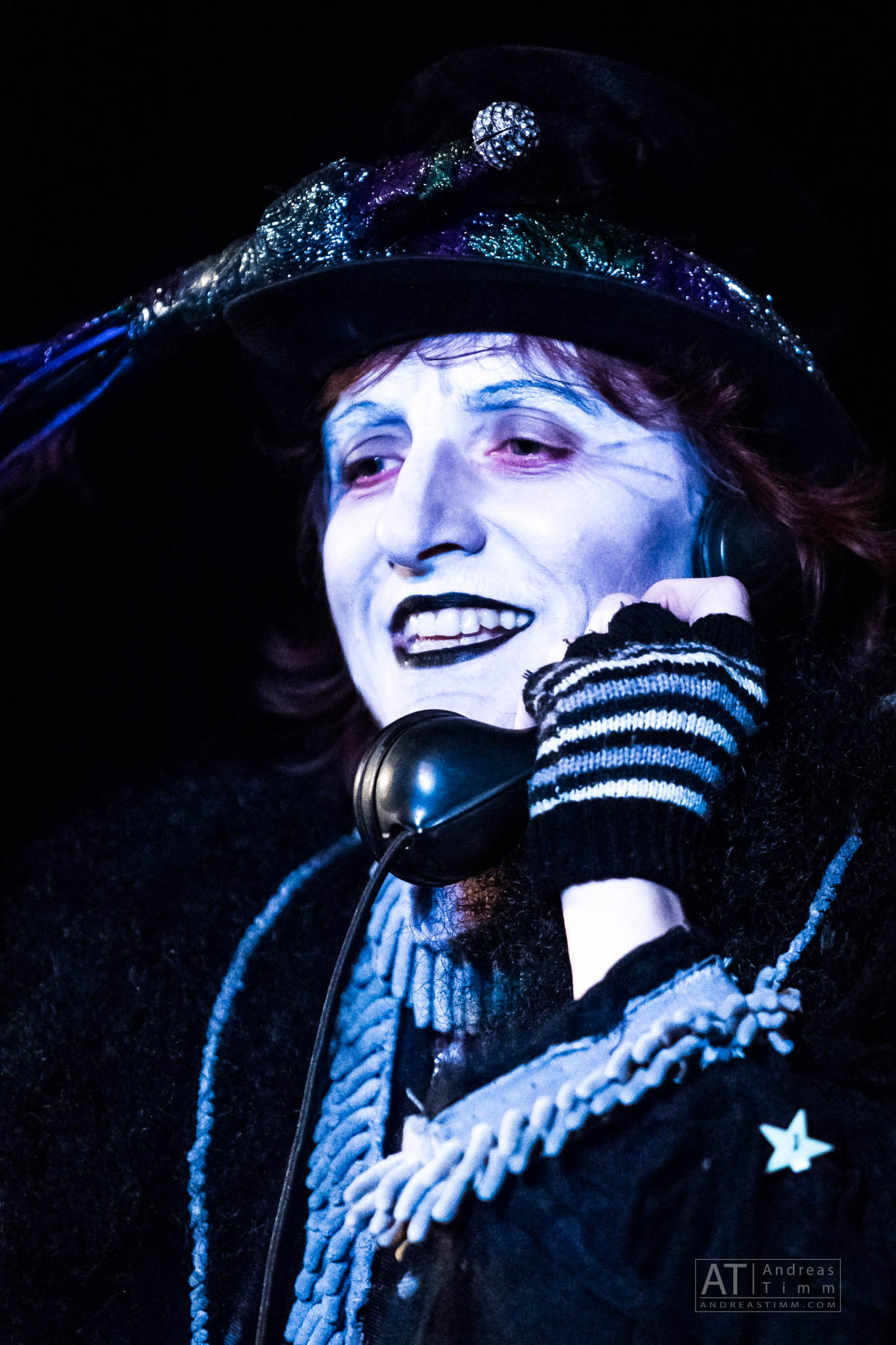 Performer in theatrical costume with white face paint talks on a vintage phone, wearing a decorated hat and striped gloves.