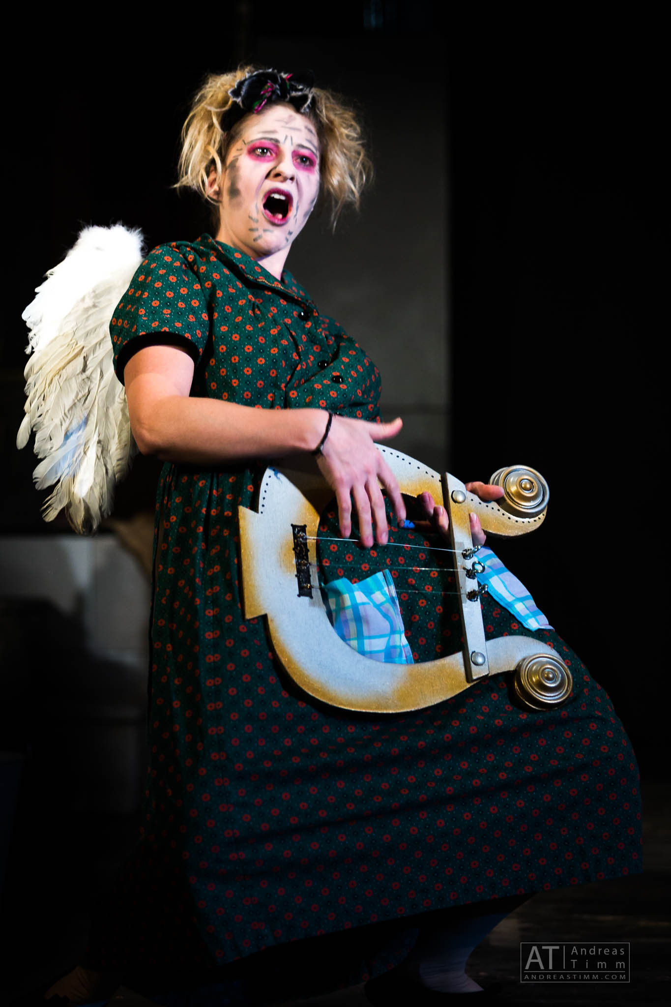 Performer in angel wings, green dress, and bold makeup, acting passionately on stage with a unique handheld prop.