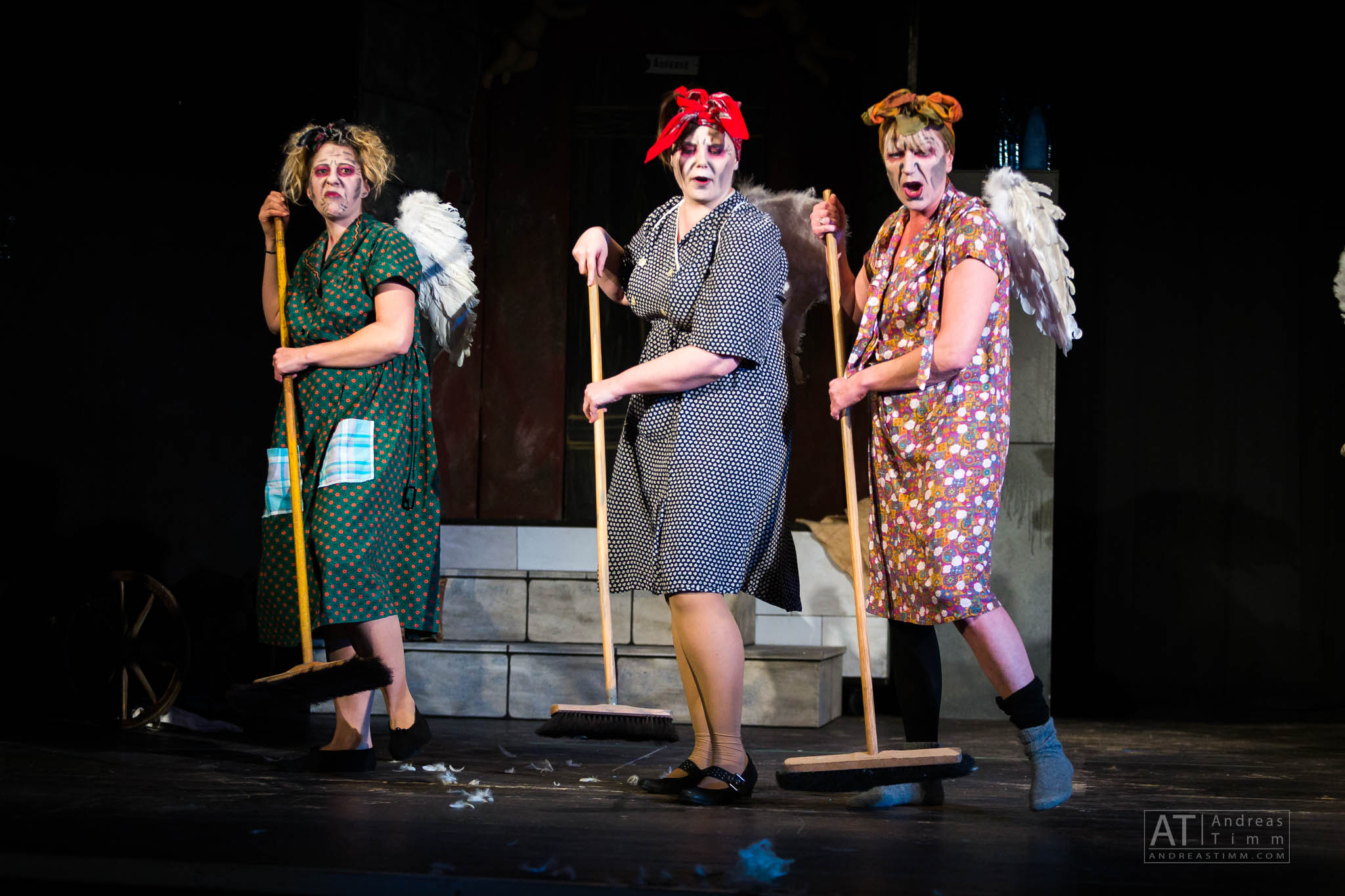 Three actors in colorful costumes and wings holding brooms on stage, performing in a theatrical comedy scene.