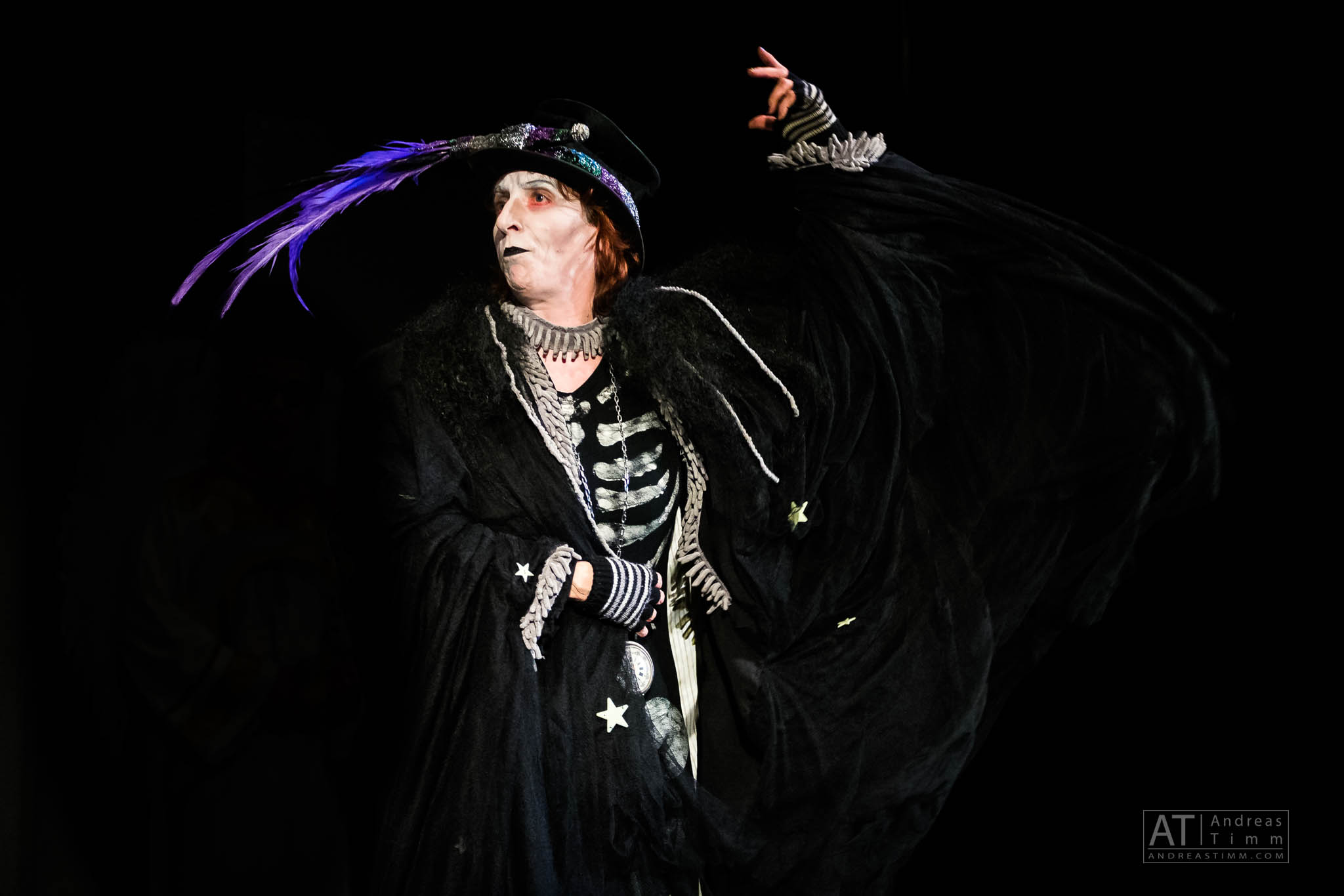 Performer in theatrical costume with a dramatic cape and feathered hat on stage, against a dark background.