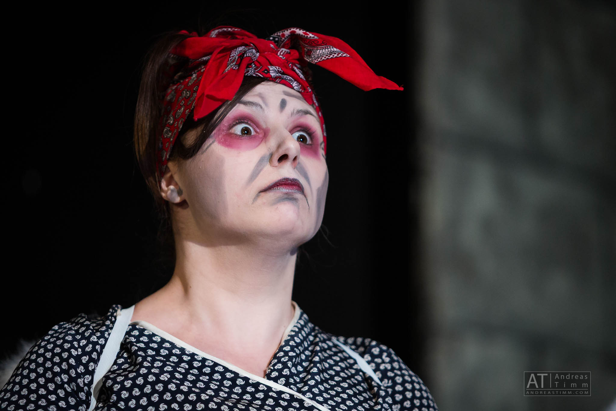 Woman in theatrical makeup and red bandana looking surprised on stage.