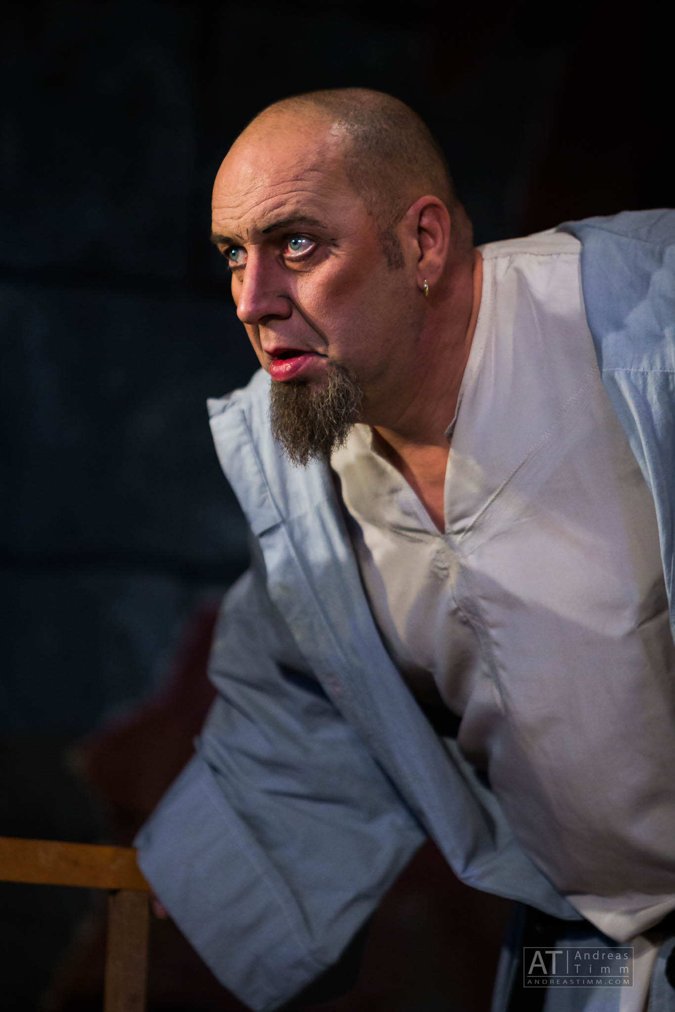 Bald man with a goatee, wearing a light shirt, looking intently off-screen in dramatic lighting.
