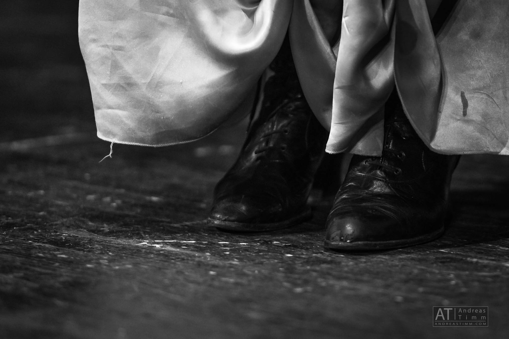 Black and white image of worn leather shoes beneath a flowing dress on a wooden floor.