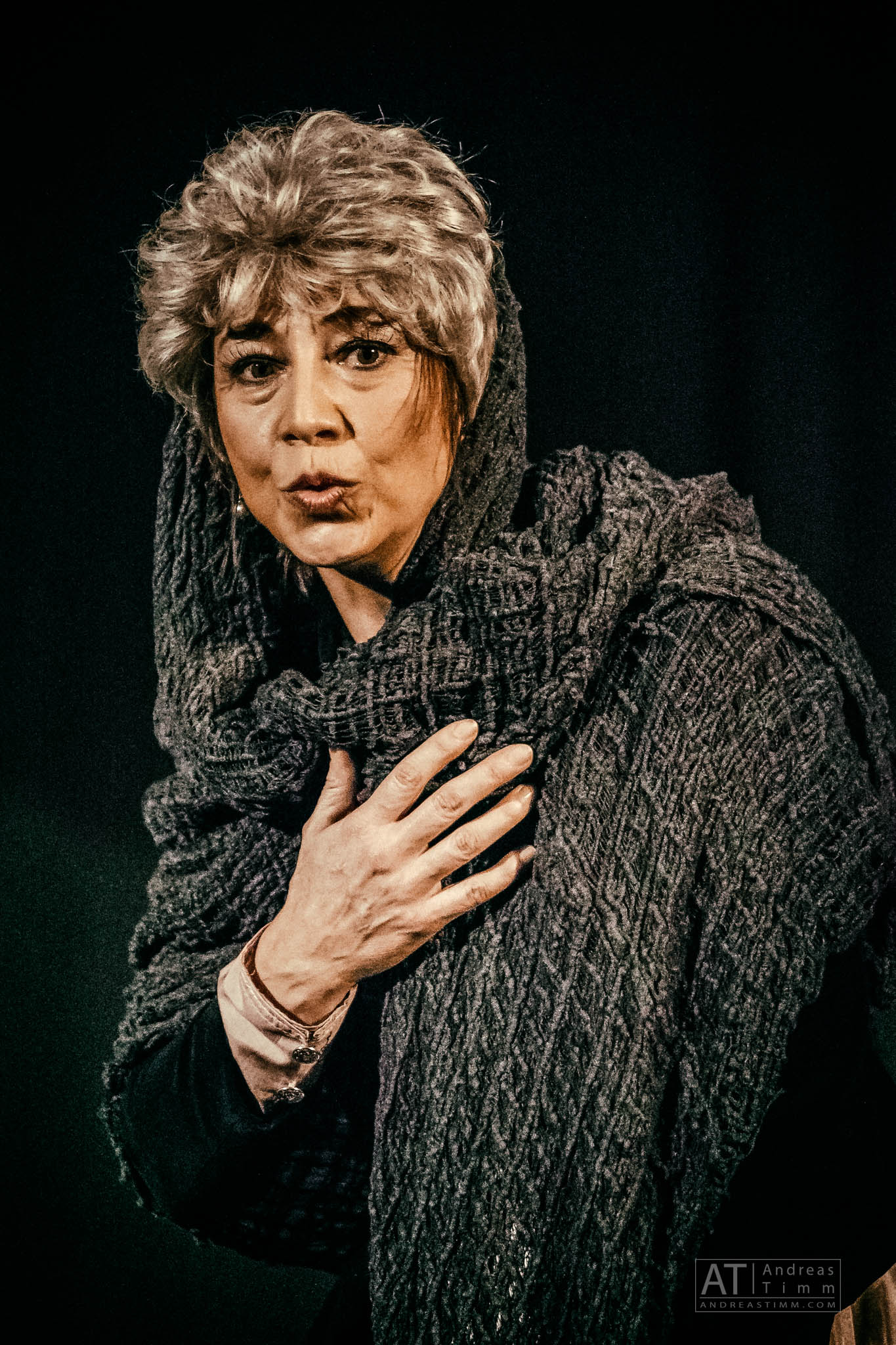 Older woman in knit shawl looks expressive on stage, hand on chest, dark background.