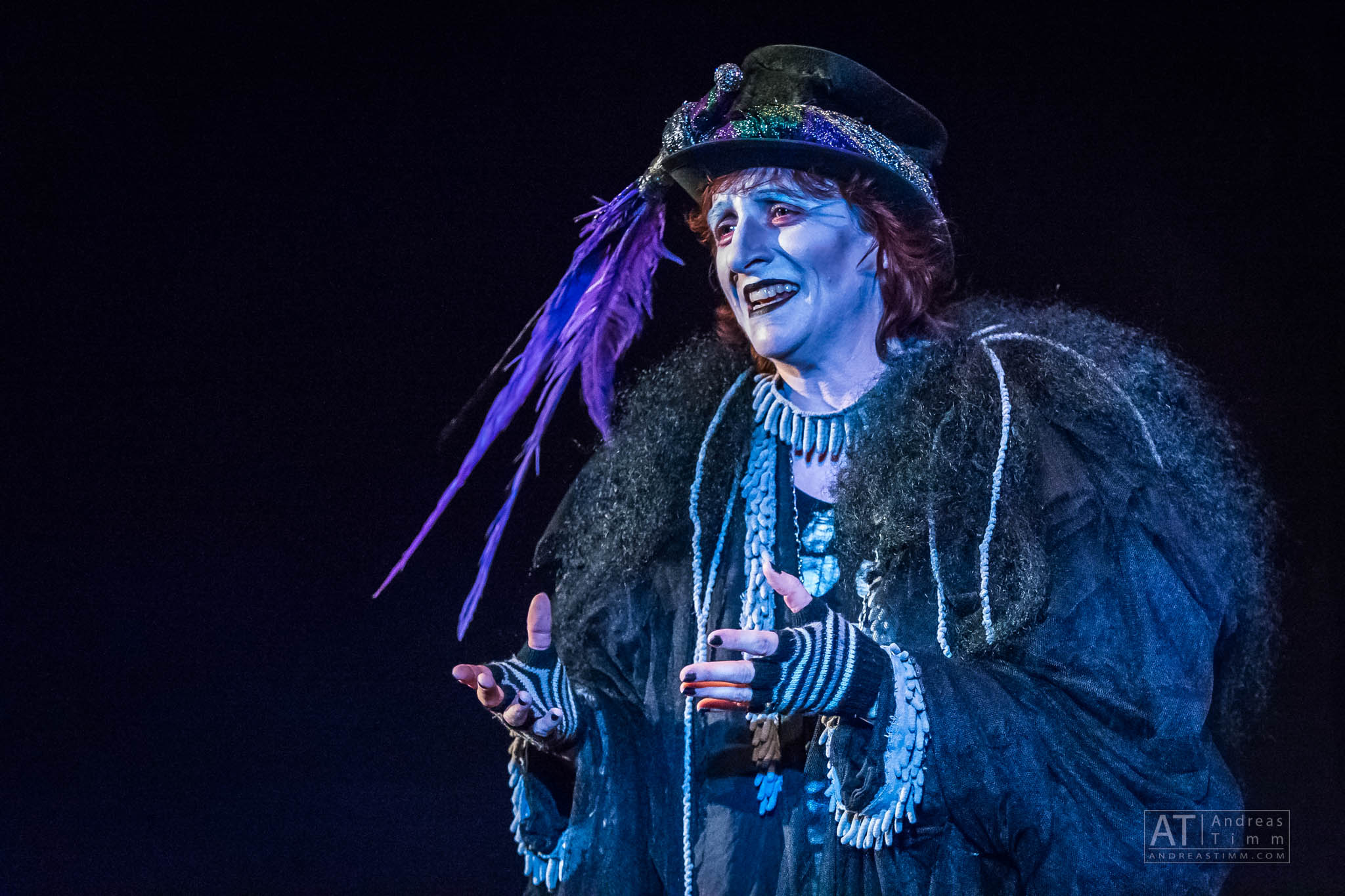 Performer in dramatic costume with colorful feathers and makeup on stage, expressing emotion.