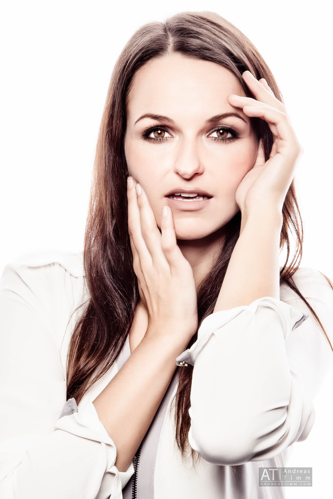 Woman with long hair in a white blouse posing with hands on face against a plain background.