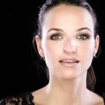 Woman with dark hair and earrings in lace top, posing against a black background with a confident expression.