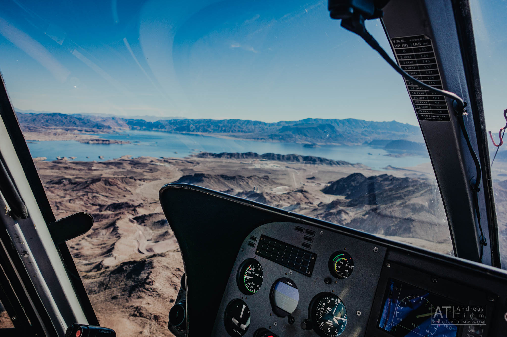 View from helicopter cockpit over desert mountains and a blue lake under clear sky.