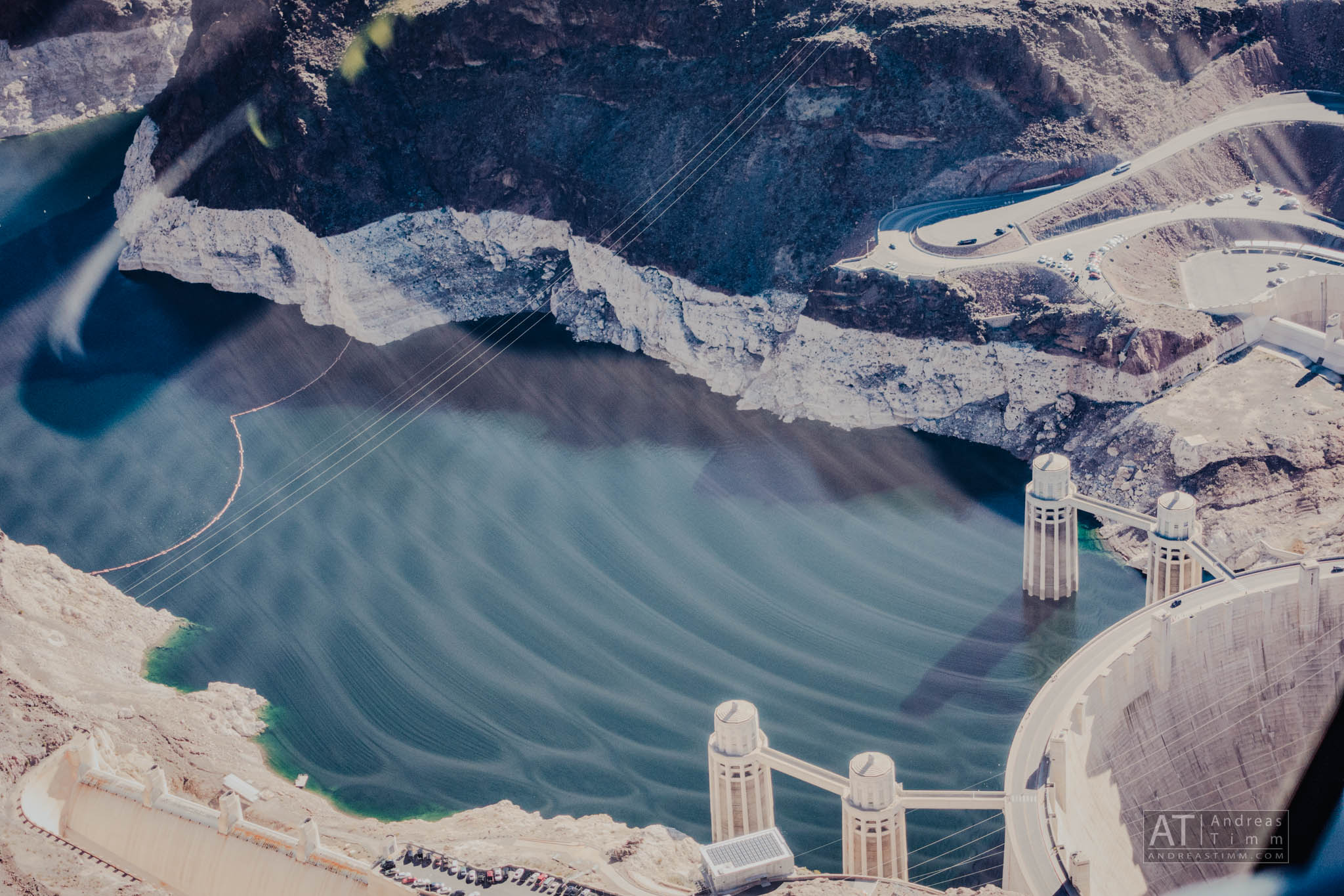 Aerial view of a large dam and reservoir surrounded by rocky terrain, with water towers and a curving road visible.