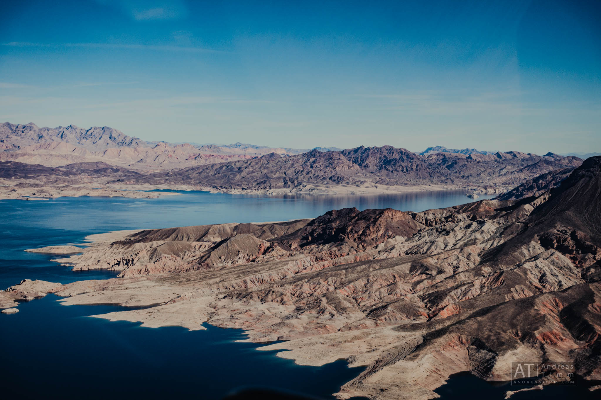Aerial view of rugged desert landscape with winding blue lake and distant mountains under a clear blue sky.