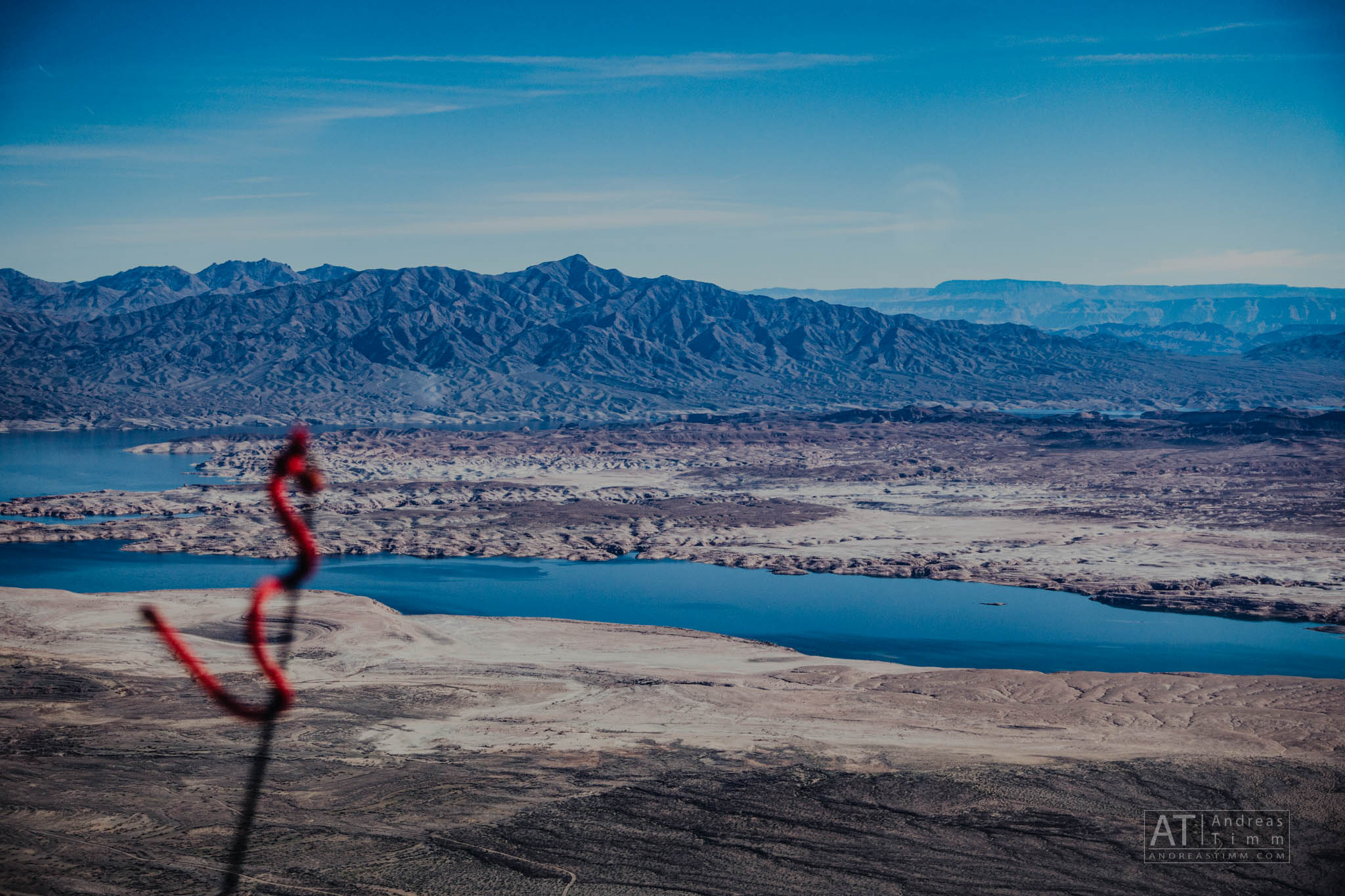 Aerial view of a desert landscape with a lake and mountains under a clear blue sky; red object blurred in the foreground.