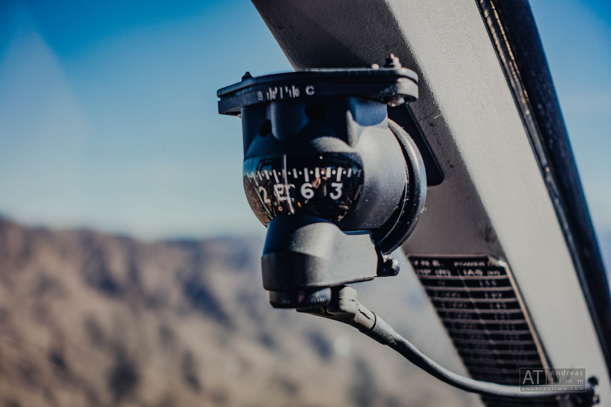 Close-up of an aircraft magnetic compass with mountainous landscape in the background under a clear blue sky.