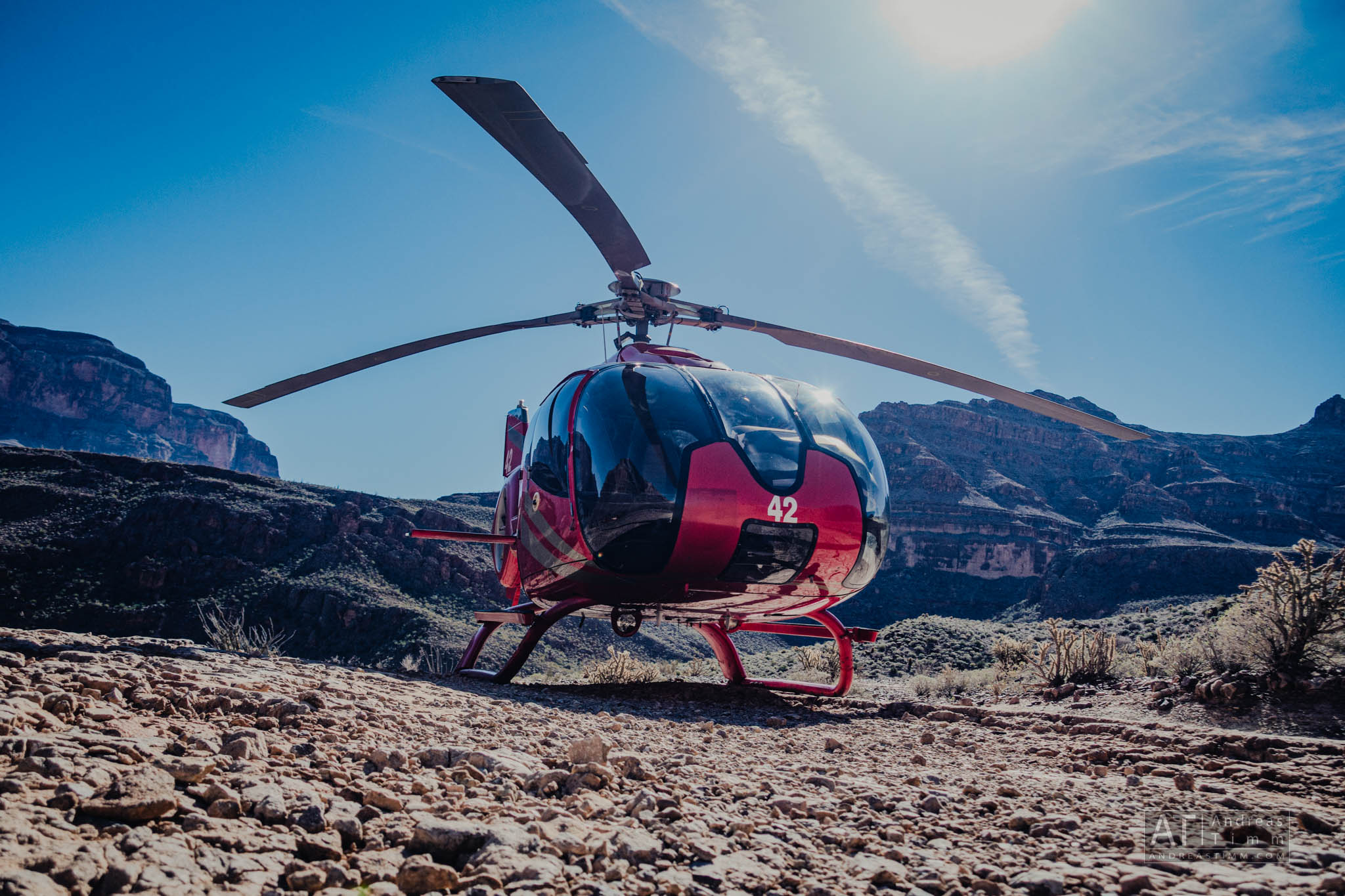 Red helicopter parked on rocky terrain with a clear blue sky and canyon mountains in the background.