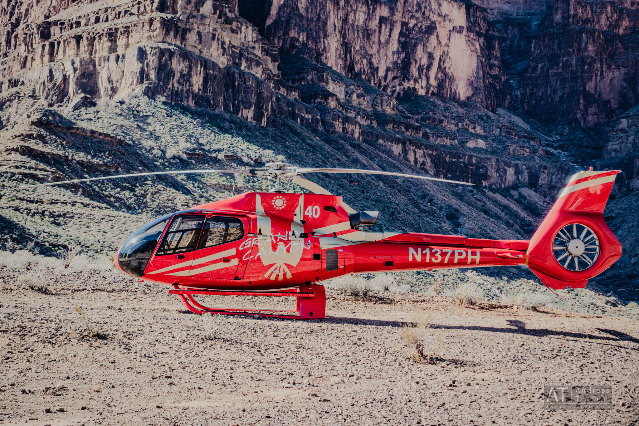 Red helicopter on rocky terrain with a canyon backdrop, labeled N137PH and Grand Canyon design on the side.