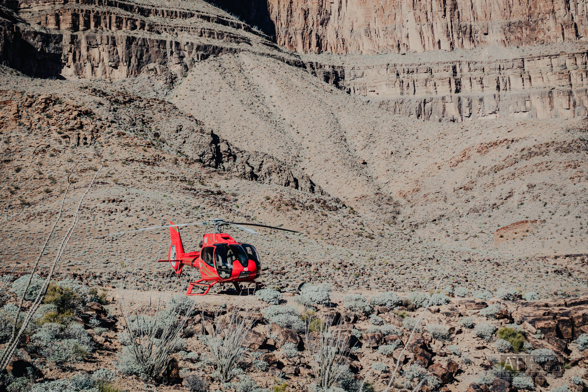 Red helicopter landed on rocky terrain in a desert canyon landscape.