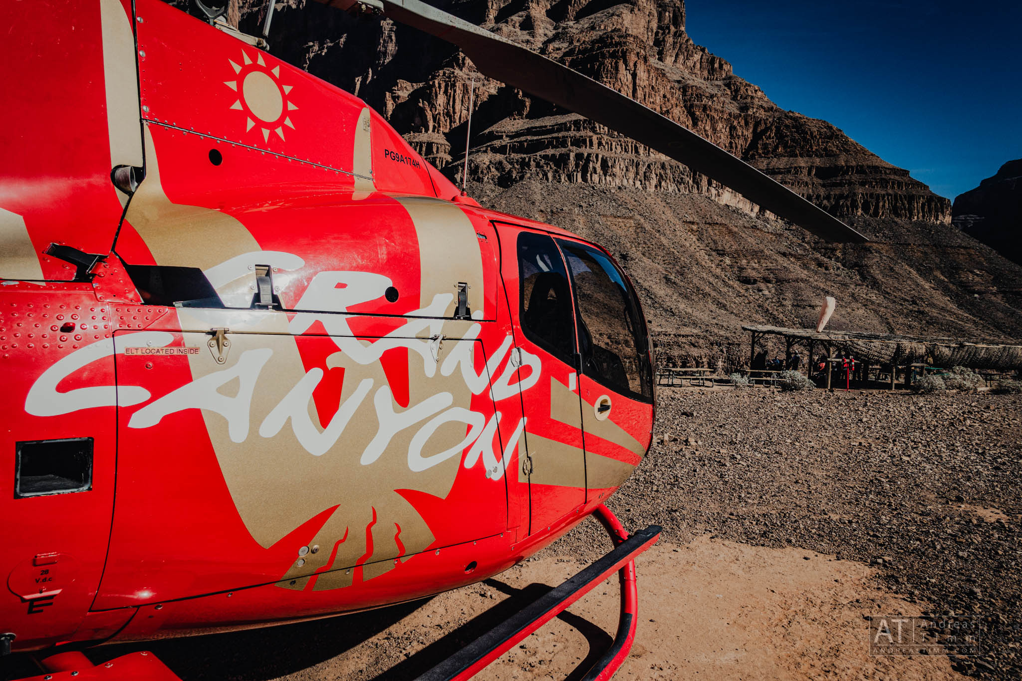 Red helicopter with Grand Canyon logo parked in rocky desert landscape under clear blue sky.
