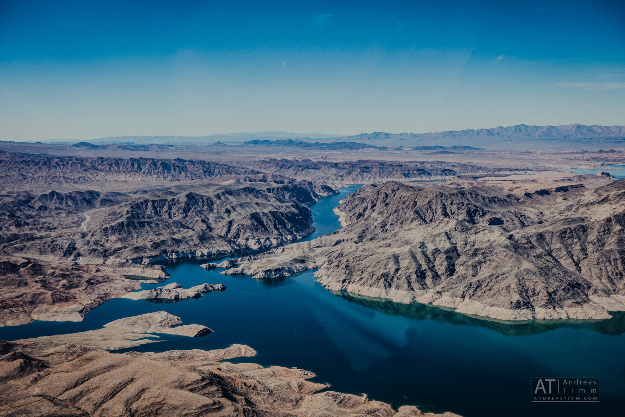 Aerial view of Lake Mead with surrounding rugged mountains and clear blue sky, showcasing desert terrain and winding water.