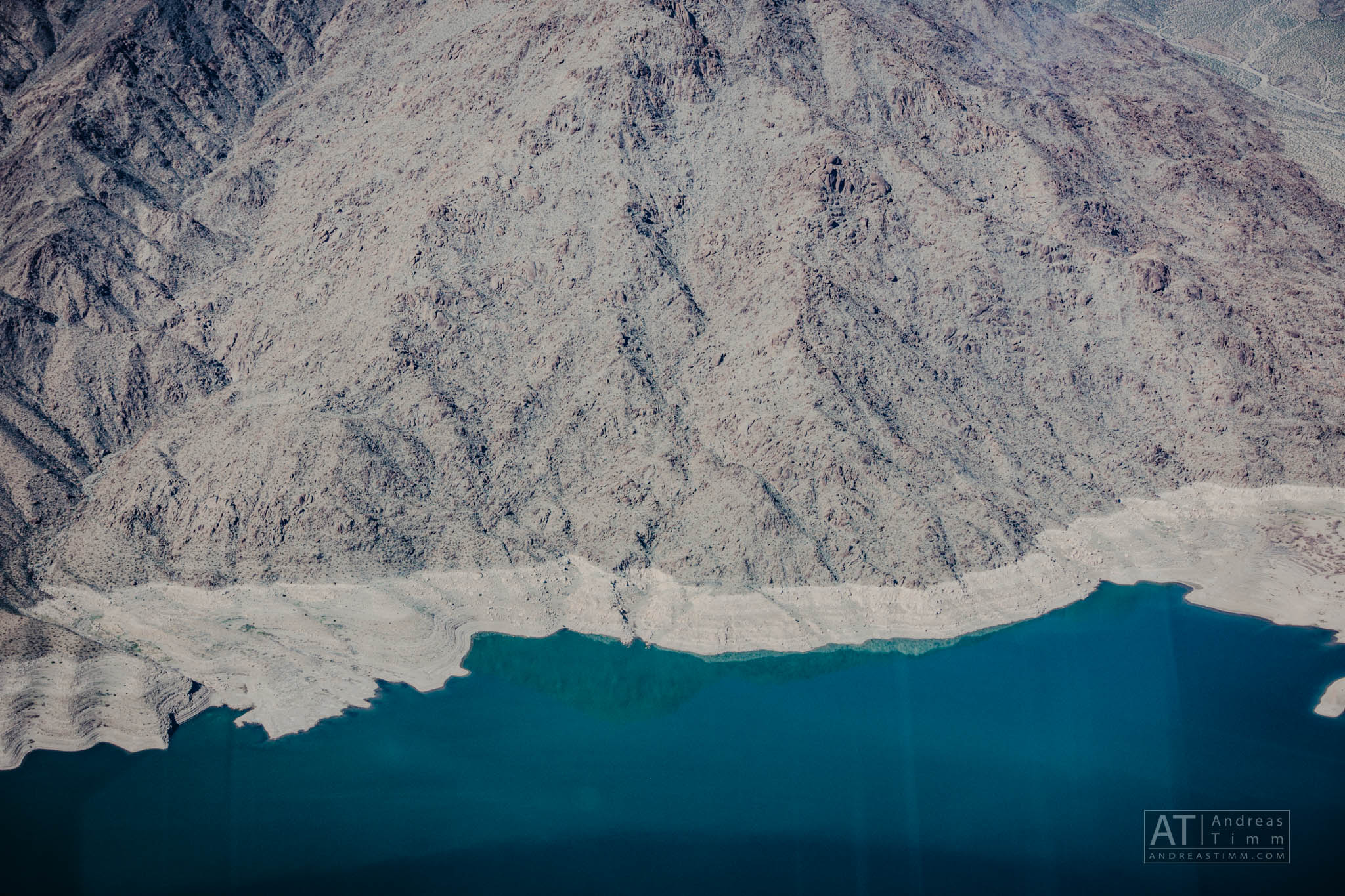 Aerial view of rugged mountain landscape meeting a calm, blue lake, showcasing the serene contrast of textures and colors.