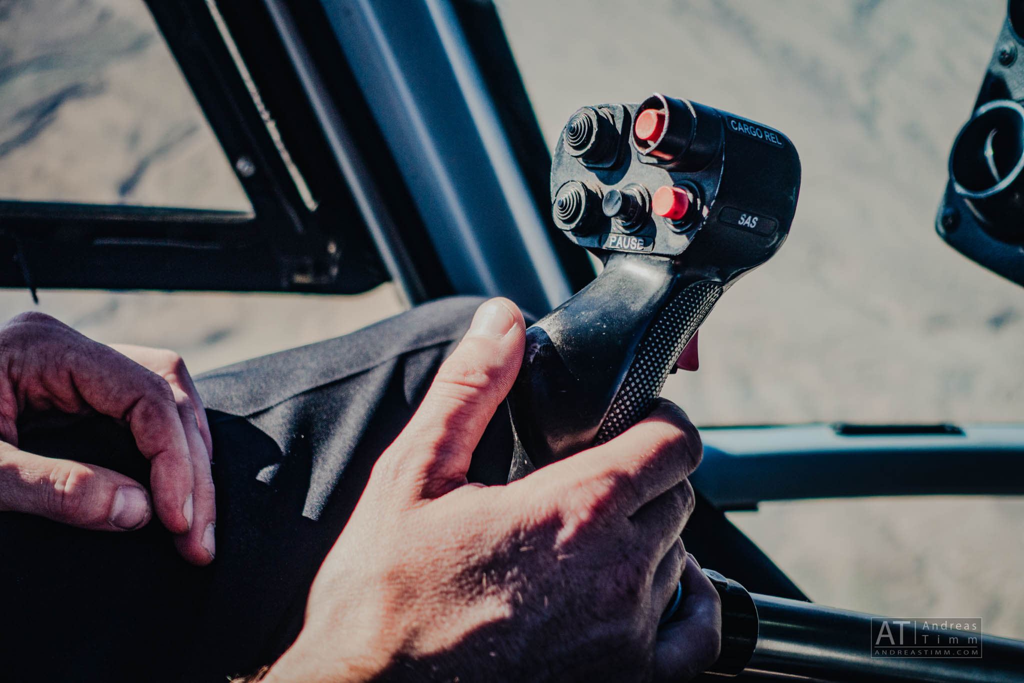 Close-up of a helicopter pilot's hands on the control stick, with visible buttons and cockpit view.