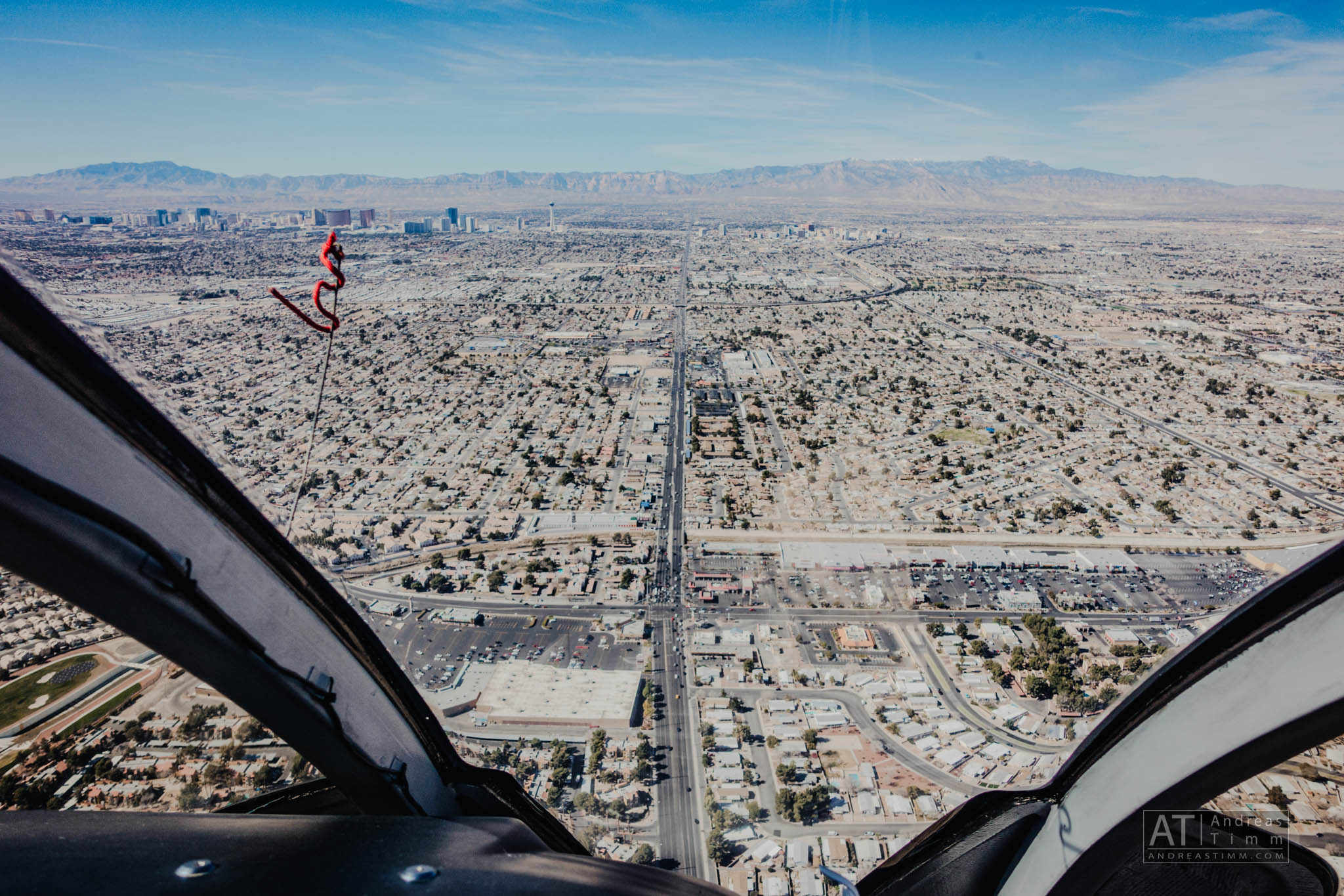 Aerial view from helicopter cockpit over a sprawling desert cityscape with distant mountains under a clear blue sky.