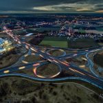Aerial view of a highway interchange at dusk with light trails, surrounded by fields and a distant town.