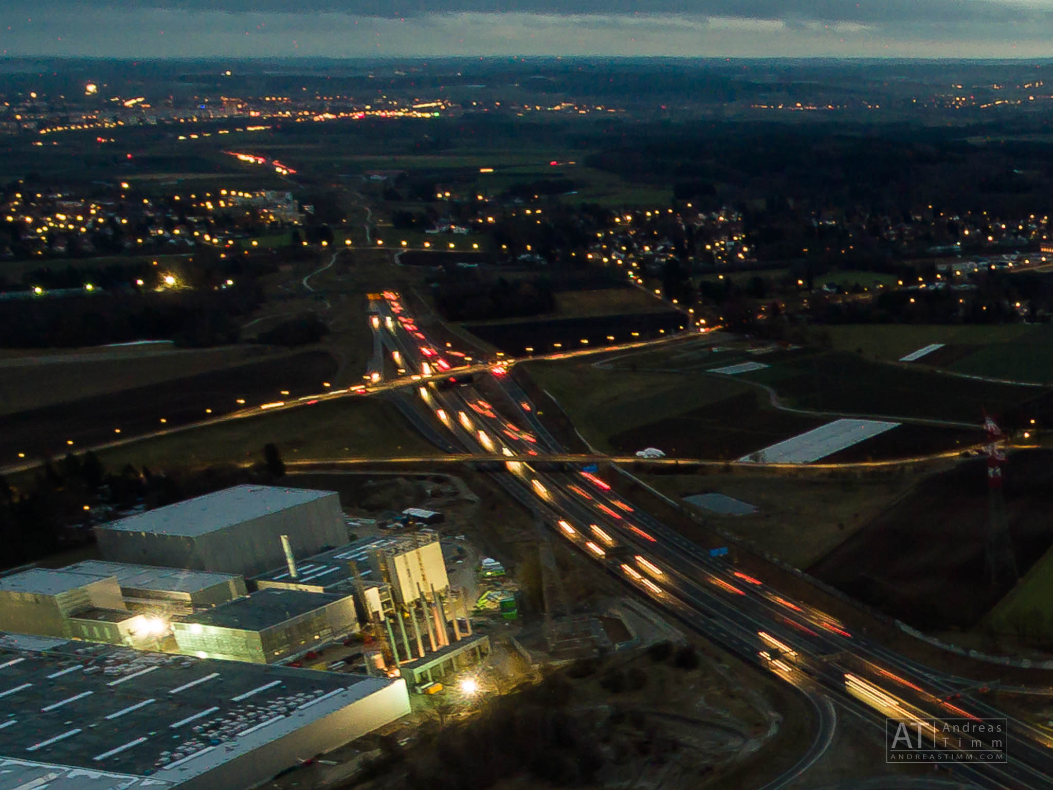 Aerial view of a highway intersection with moving traffic lights, industrial buildings, and rural landscape at dusk.