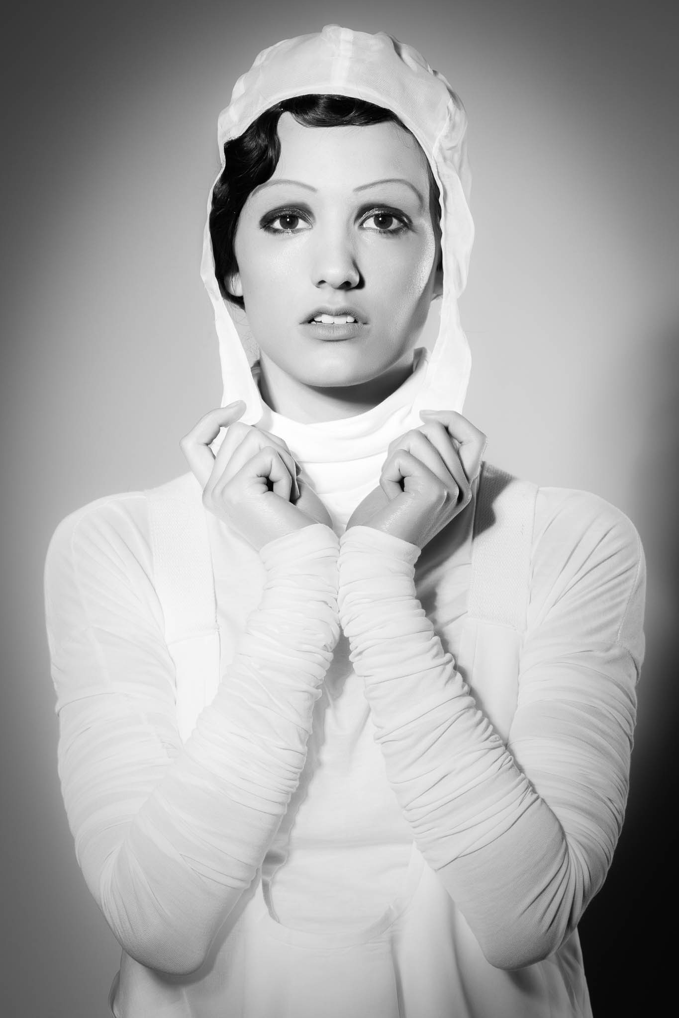 Black and white photo of a woman in vintage attire, wearing a white hooded outfit and staring intently at the camera.