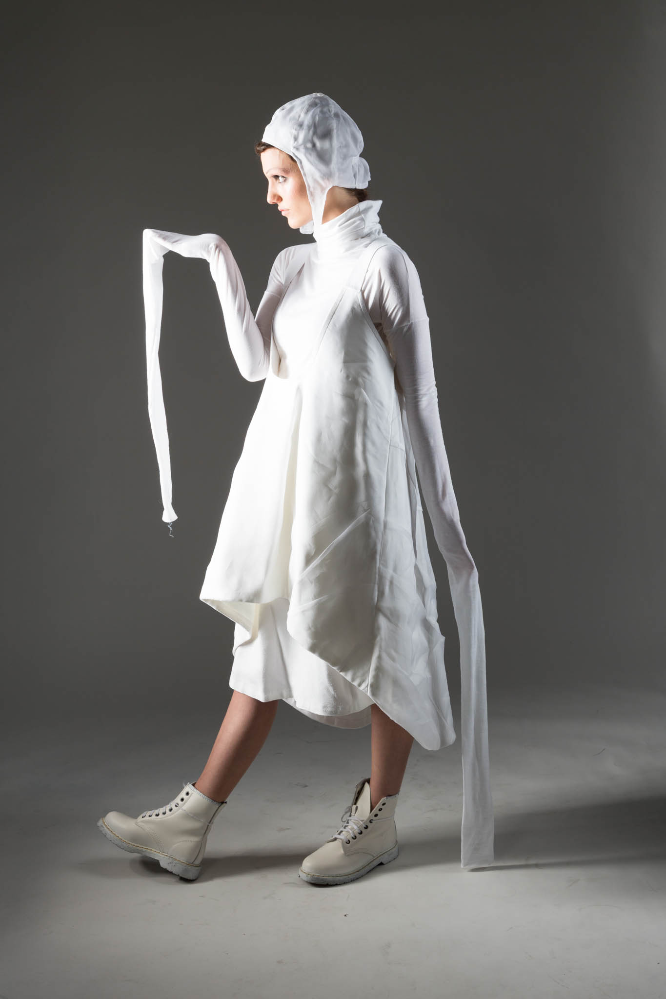 Avant-garde fashion model in white asymmetrical dress and boots, striking a side pose on a gray background.