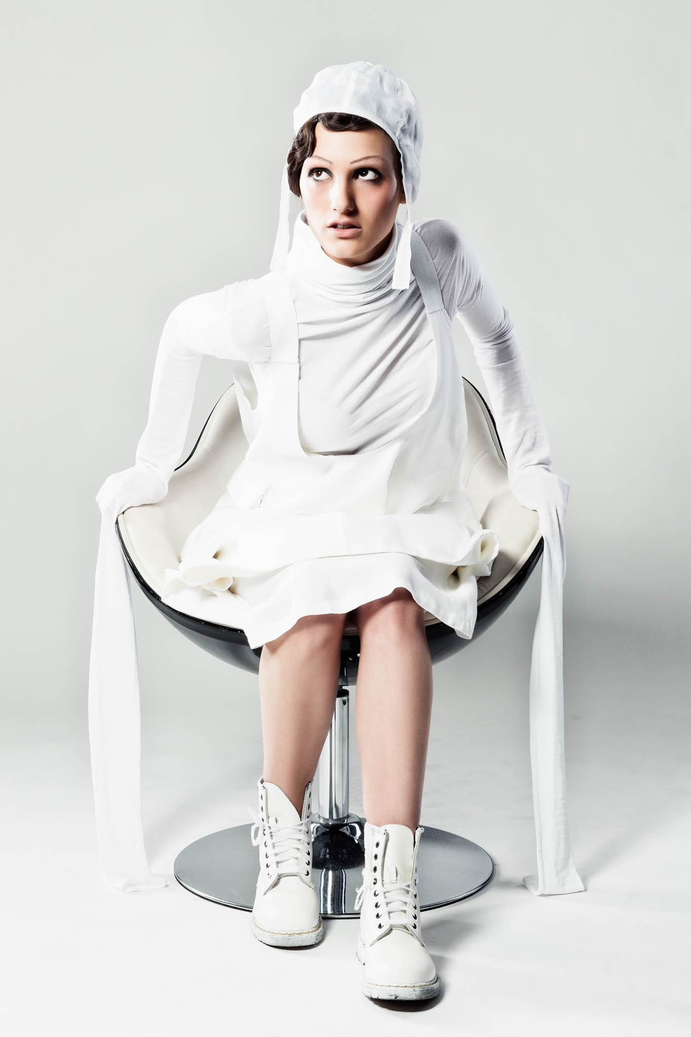 Woman in white avant-garde outfit sits on modern chair, wearing boots and a cap, posing against a plain background.