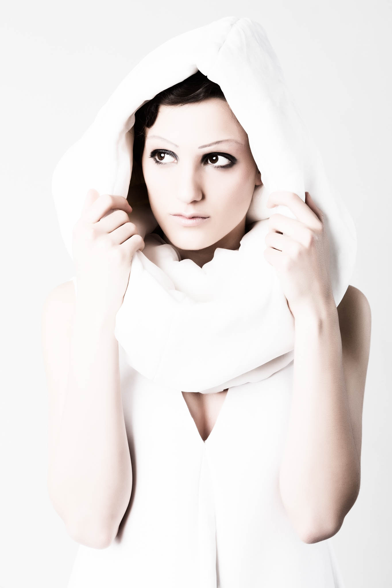 Modern woman in white cloak with hood, striking makeup, gazing sideways against a bright background.