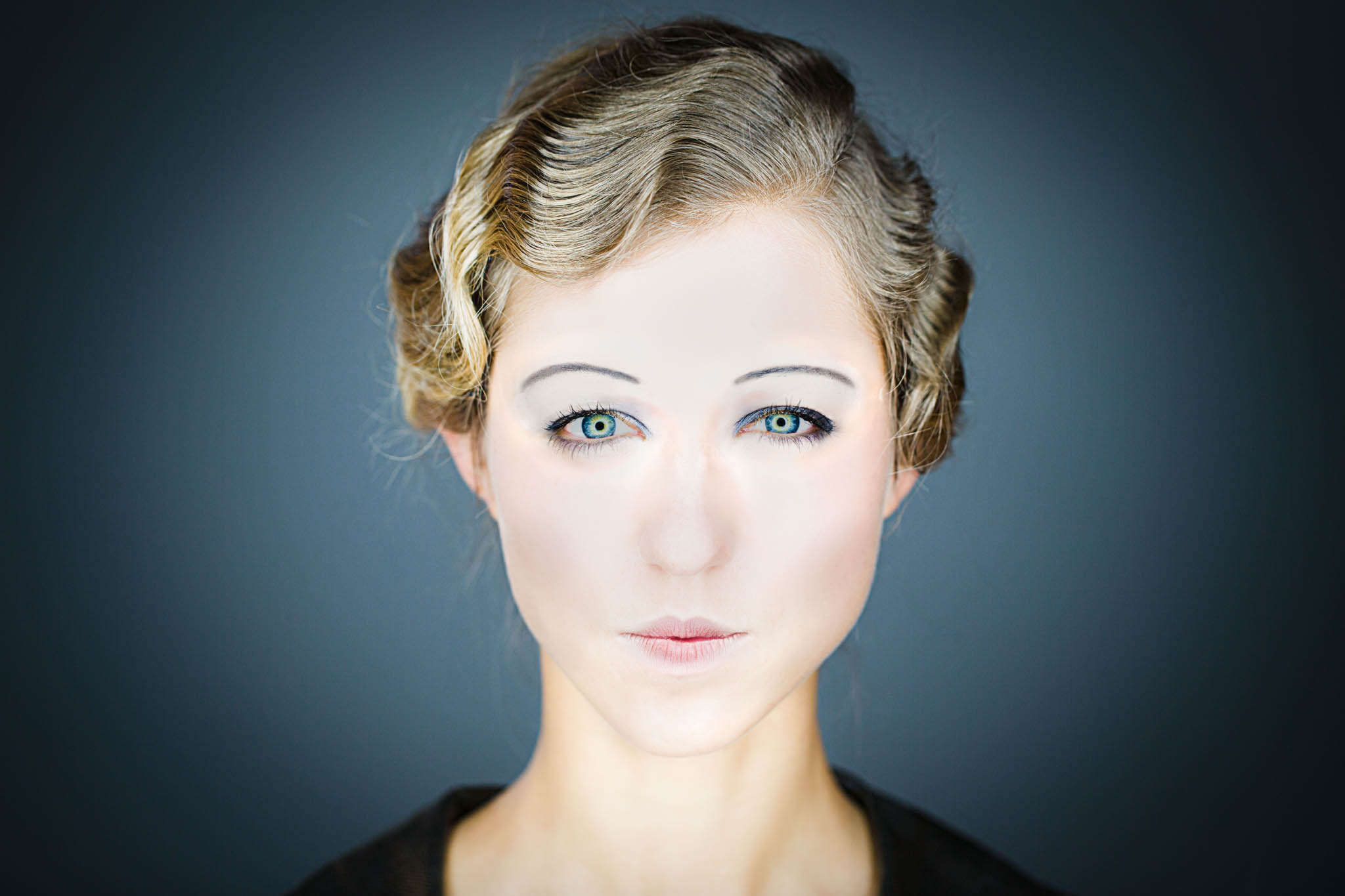 Close-up of a person with vintage hairstyle, smooth complexion, and bright eyes against a dark background.