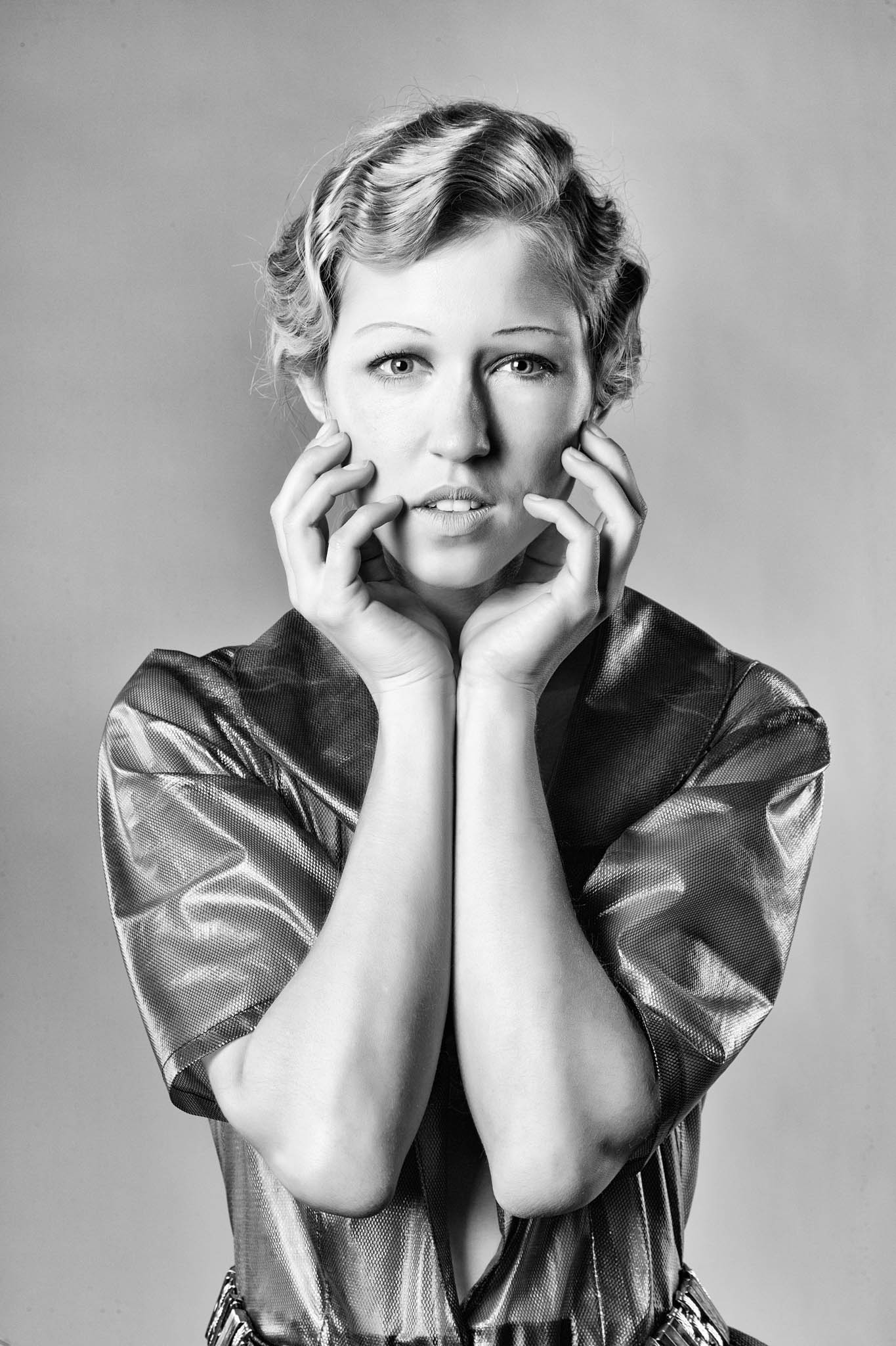 Black and white portrait of a woman with wavy hair, hands near face, wearing a shiny textured top.