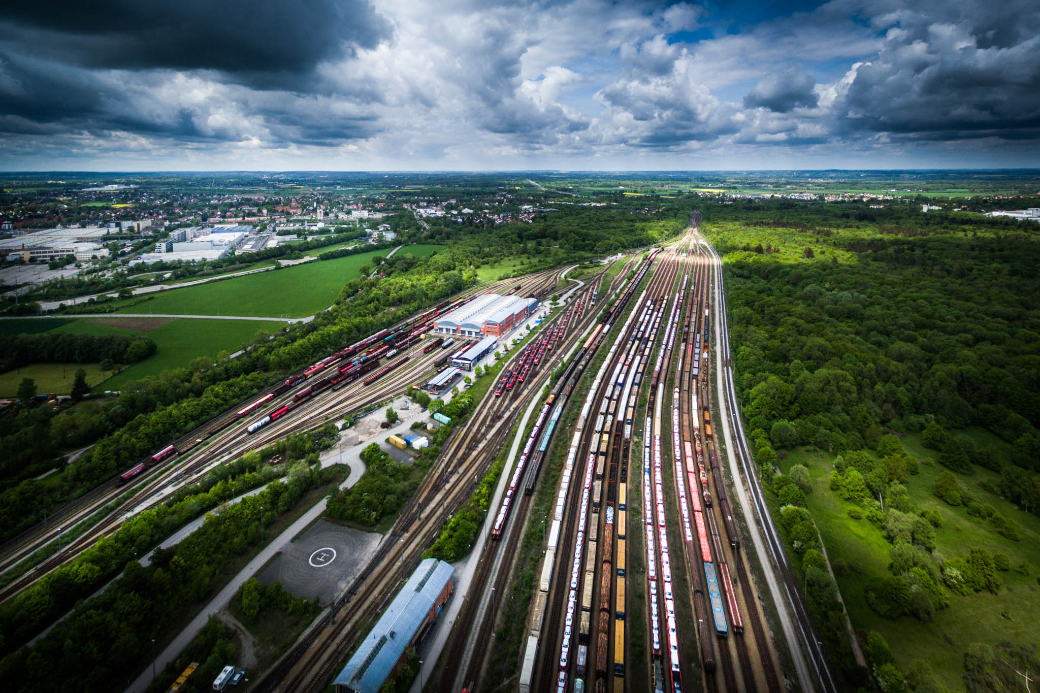 Aerial view of a bustling railyard with numerous trains amid lush green fields and a dramatic cloudy sky.
