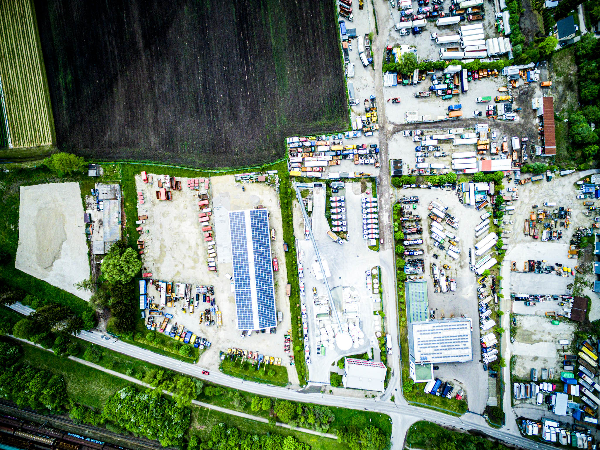 Aerial view of industrial area with parked trucks, solar panels on roofs, and surrounding greenery.