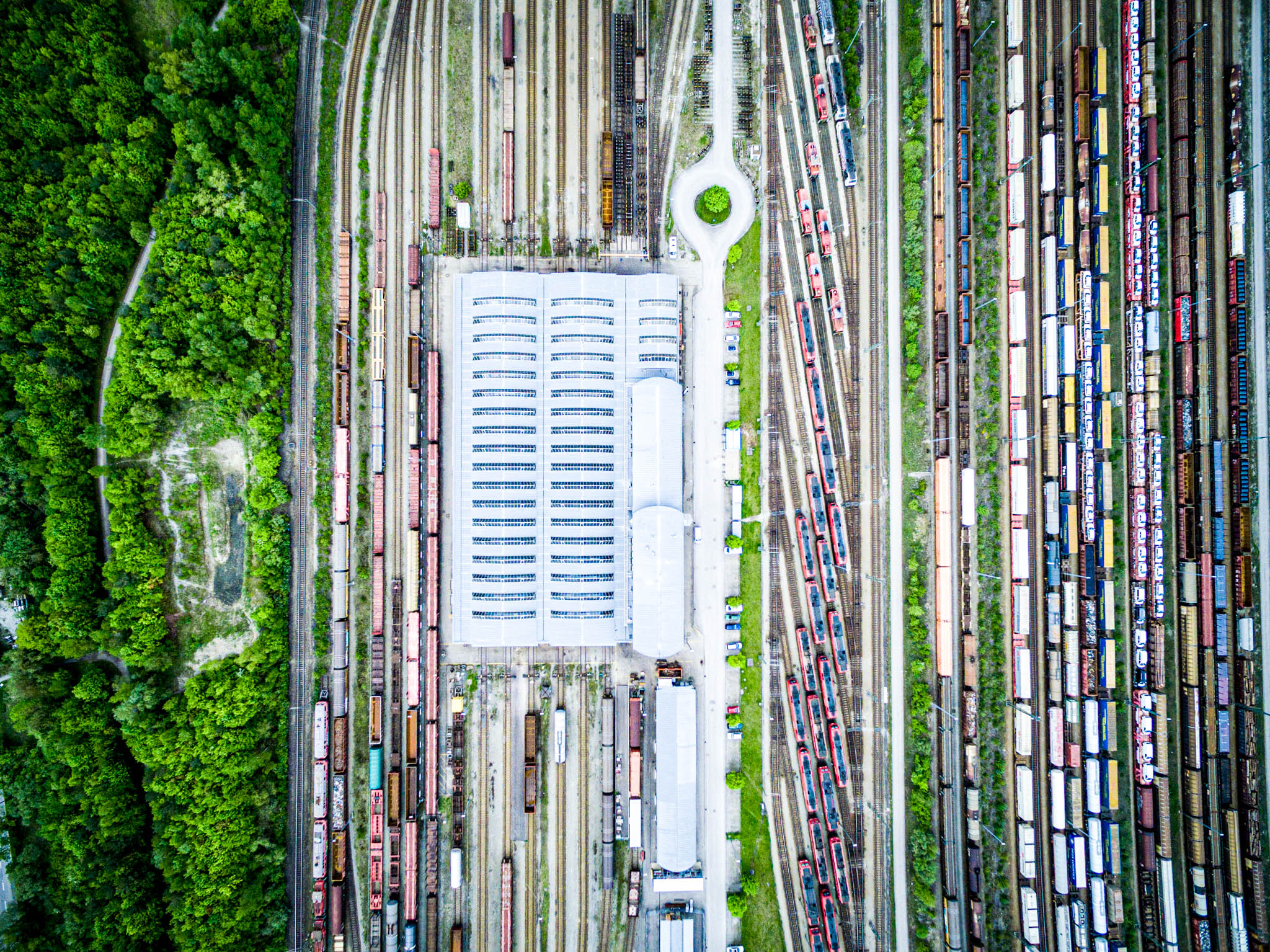 Aerial view of a train depot with multiple rail tracks and trains, bordered by dense green forest on the left.