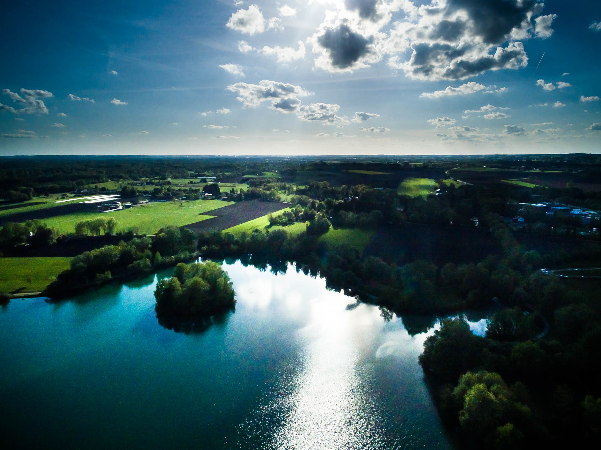 Aerial view of a serene lake reflecting clouds, surrounded by lush green fields and trees under a partly cloudy sky.