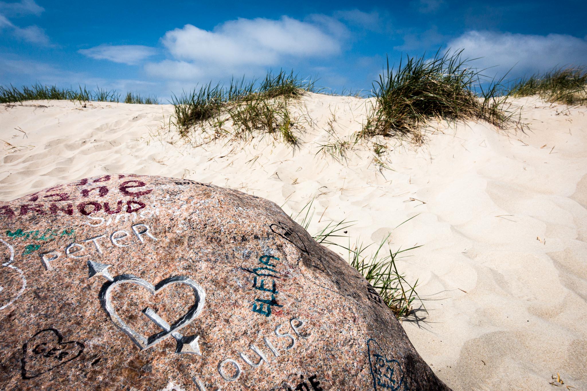 Rock with carved love heart and names on a sandy beach with dune grasses under a blue sky.