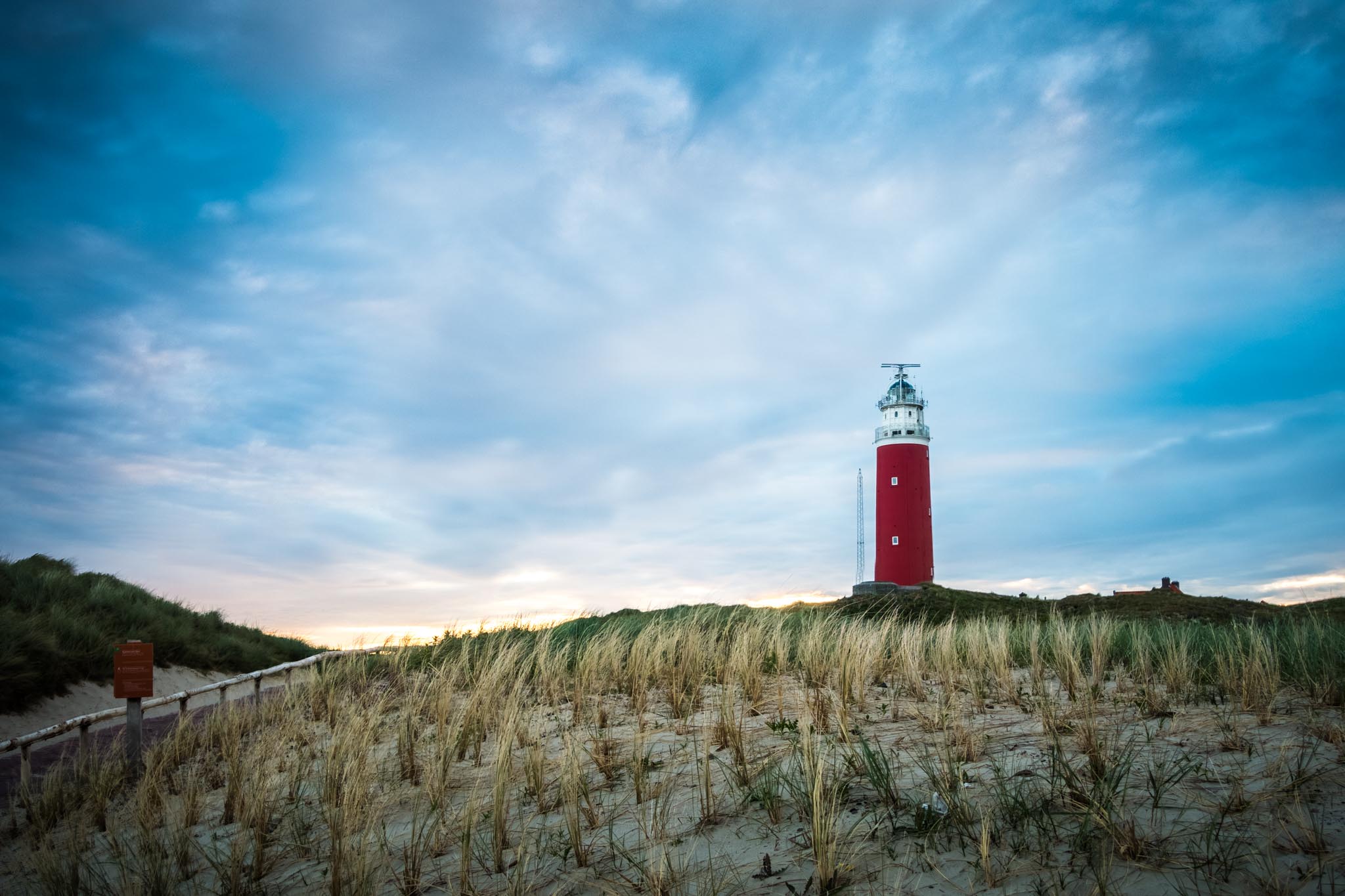 Red lighthouse on a sandy hill with grassy dunes at sunset under a cloudy blue sky.