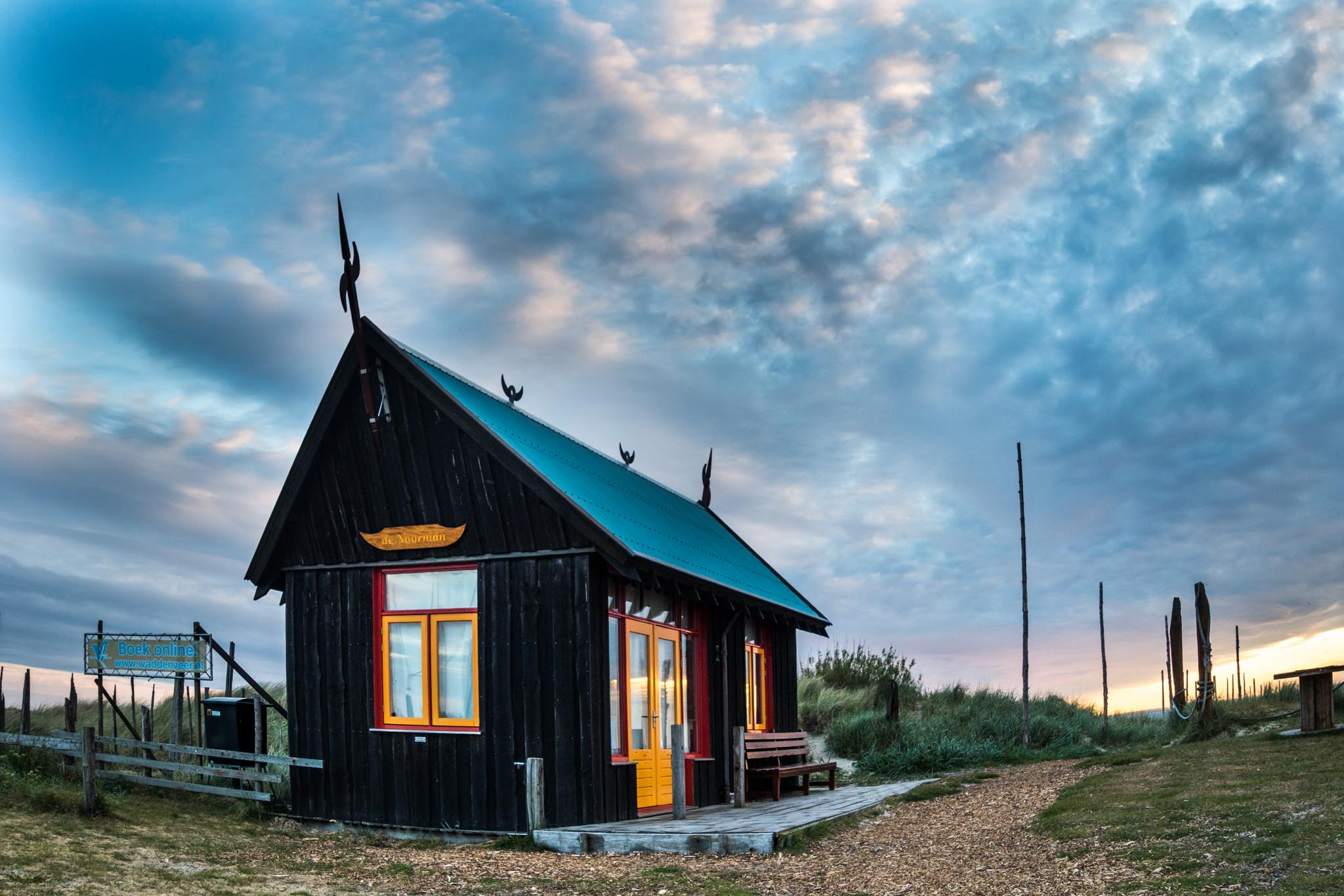 Rustic wooden cabin with yellow-trimmed windows under a dramatic cloudy sky, surrounded by grass and wooden poles.