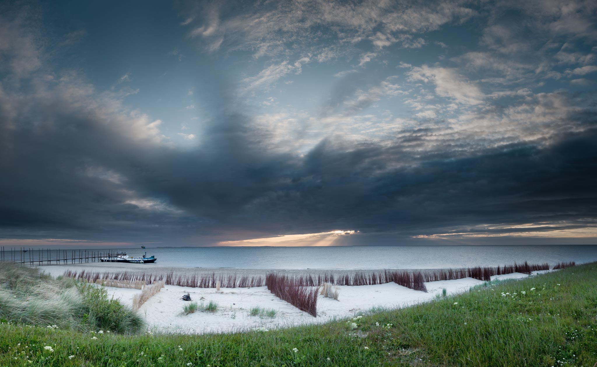 Serene beach at sunset with grassy dunes, dark clouds, and a lone boat on the calm sea.