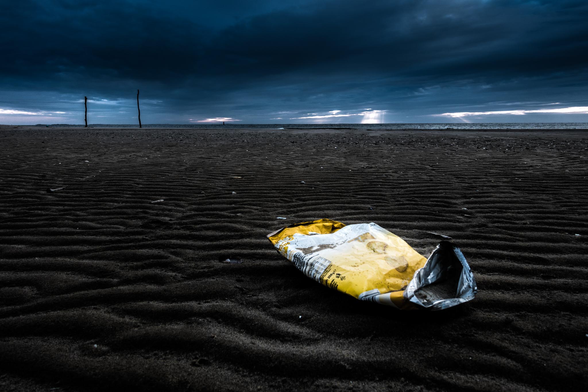 Discarded snack bag on a cloudy beach at dusk, with sand ripples and distant ocean view.