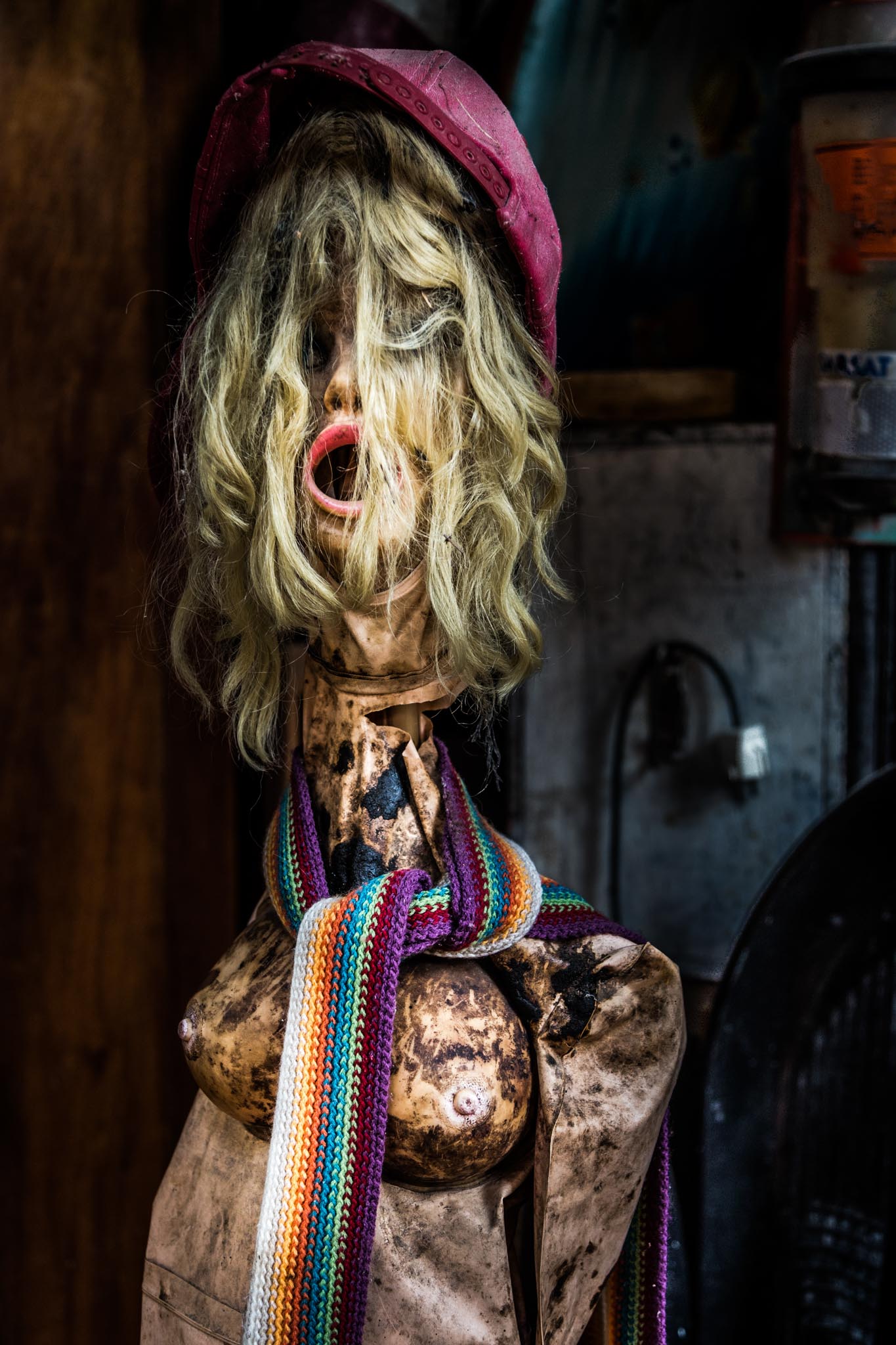 Distressed mannequin with blond wig, red hat, and colorful scarf, set against a dark, rustic background.