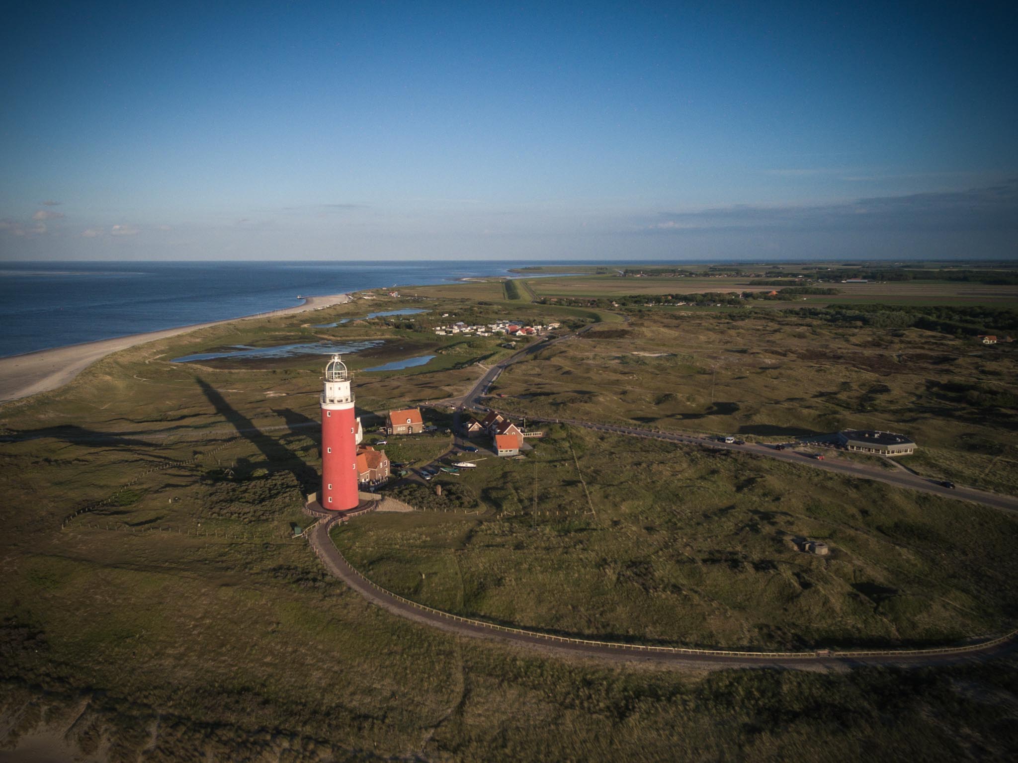 Red lighthouse on a coastal landscape with sandy beach and grassy fields, viewed from above under a clear blue sky.