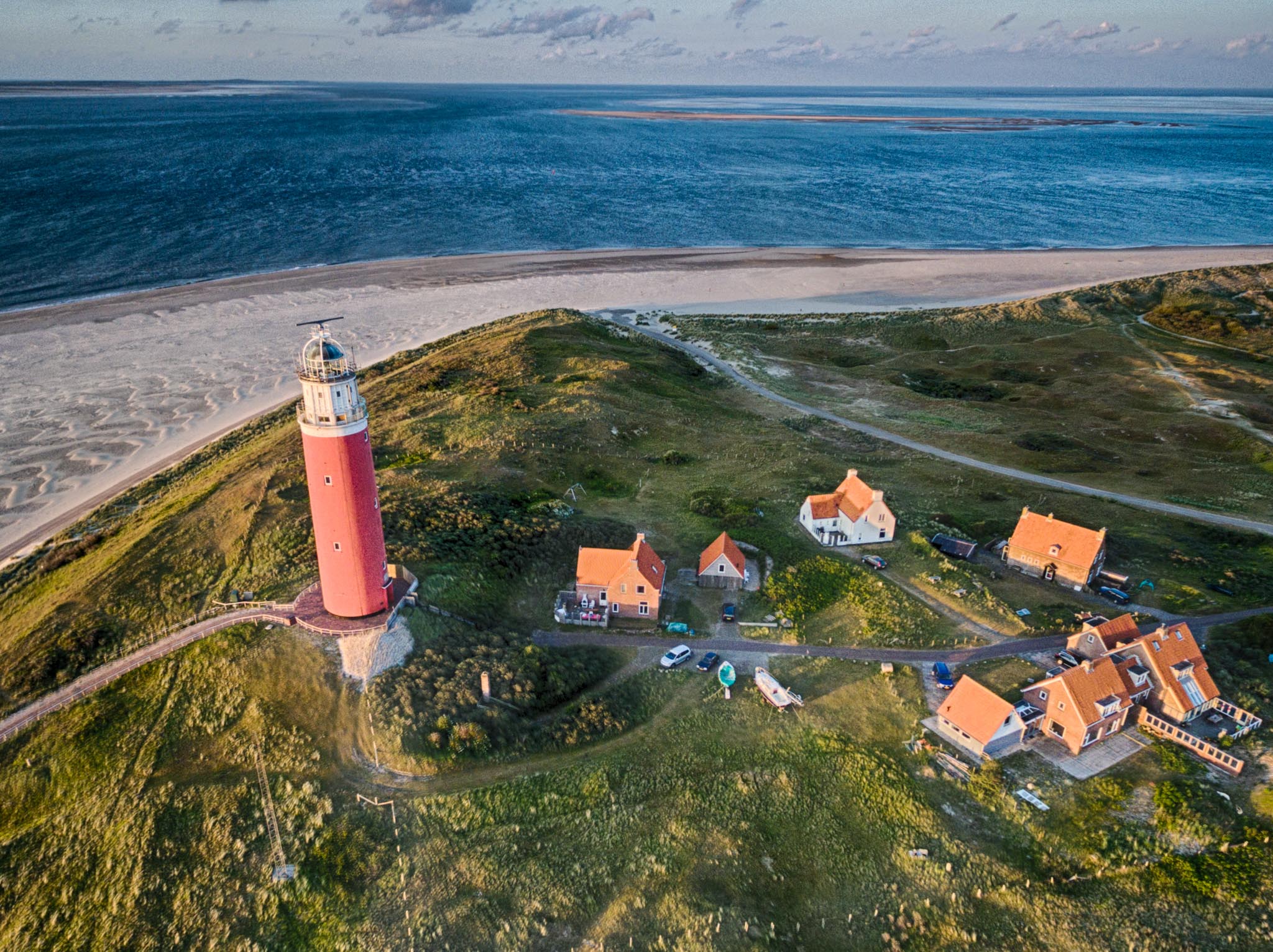 Aerial view of red lighthouse and nearby orange-roofed houses on green hills by the sea and sandy beach.
