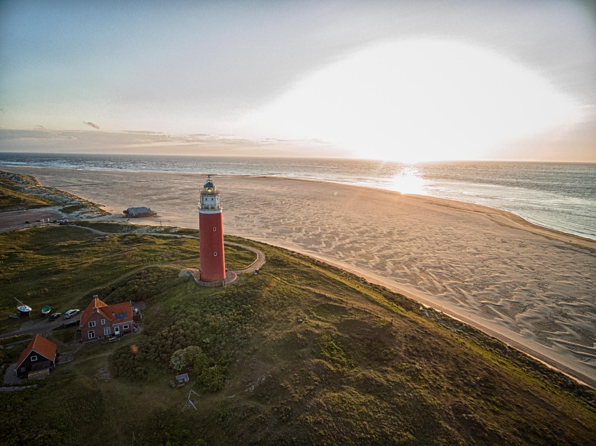 Aerial view of a red lighthouse on a grassy hill by a sandy beach at sunset, with ocean waves in the background.