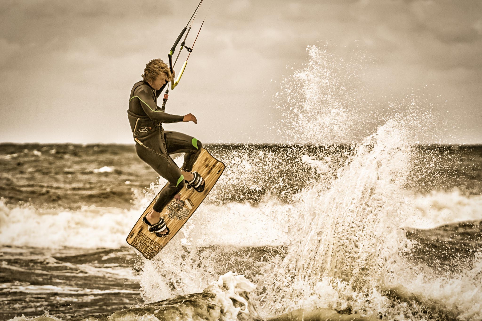 Kitesurfer performs jump over waves on ocean, creating splash; overcast sky sets dramatic tone.