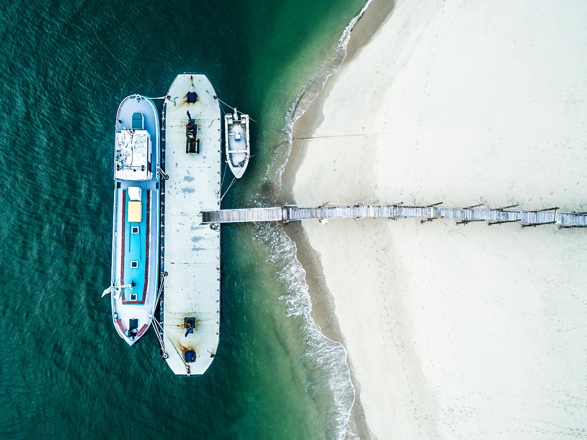 Aerial view of a blue boat docked at a wooden pier beside a sandy beach with gentle waves.