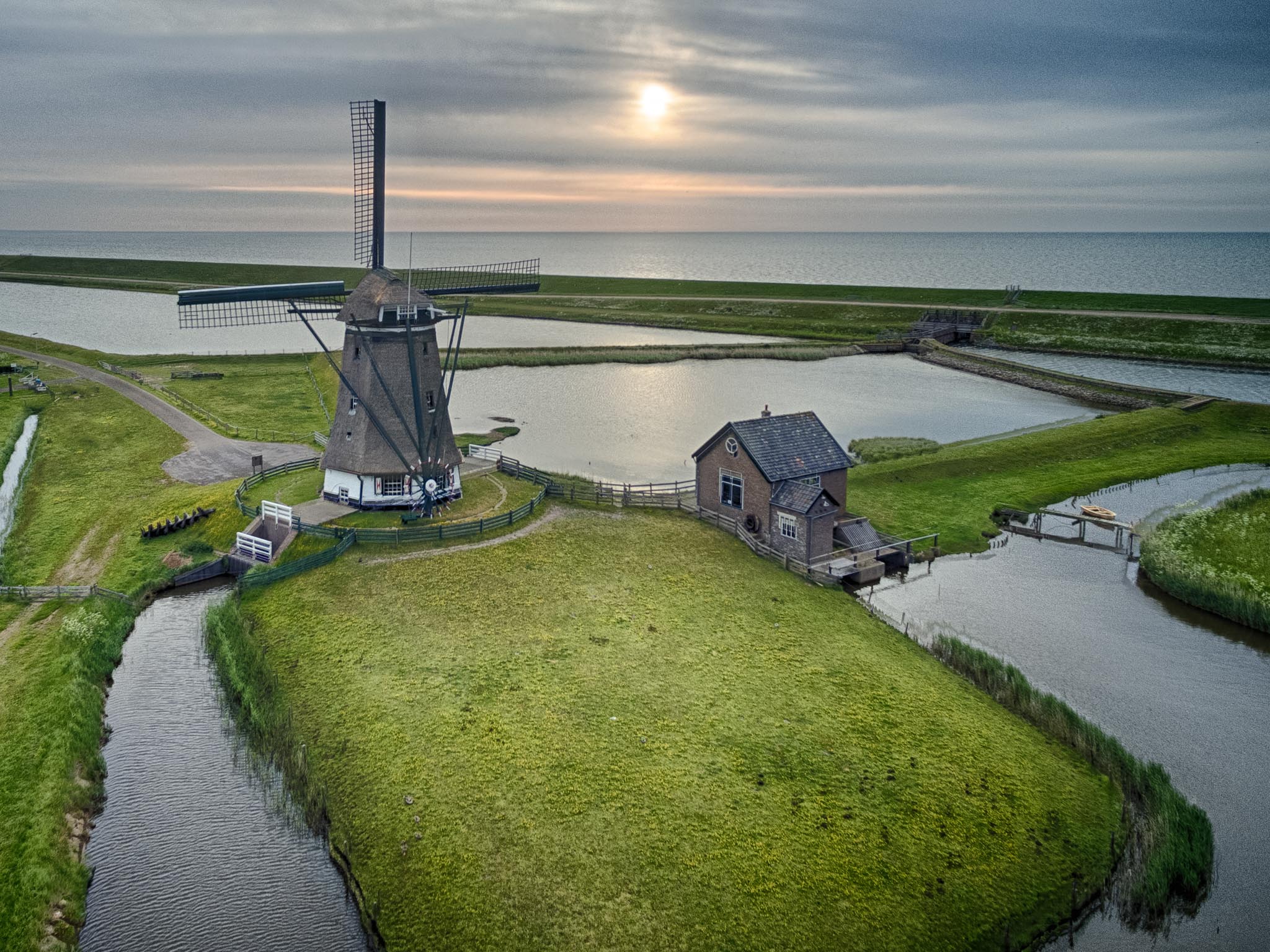Aerial view of a windmill by the coast with surrounding waterways and a small building under a cloudy sky at sunset.